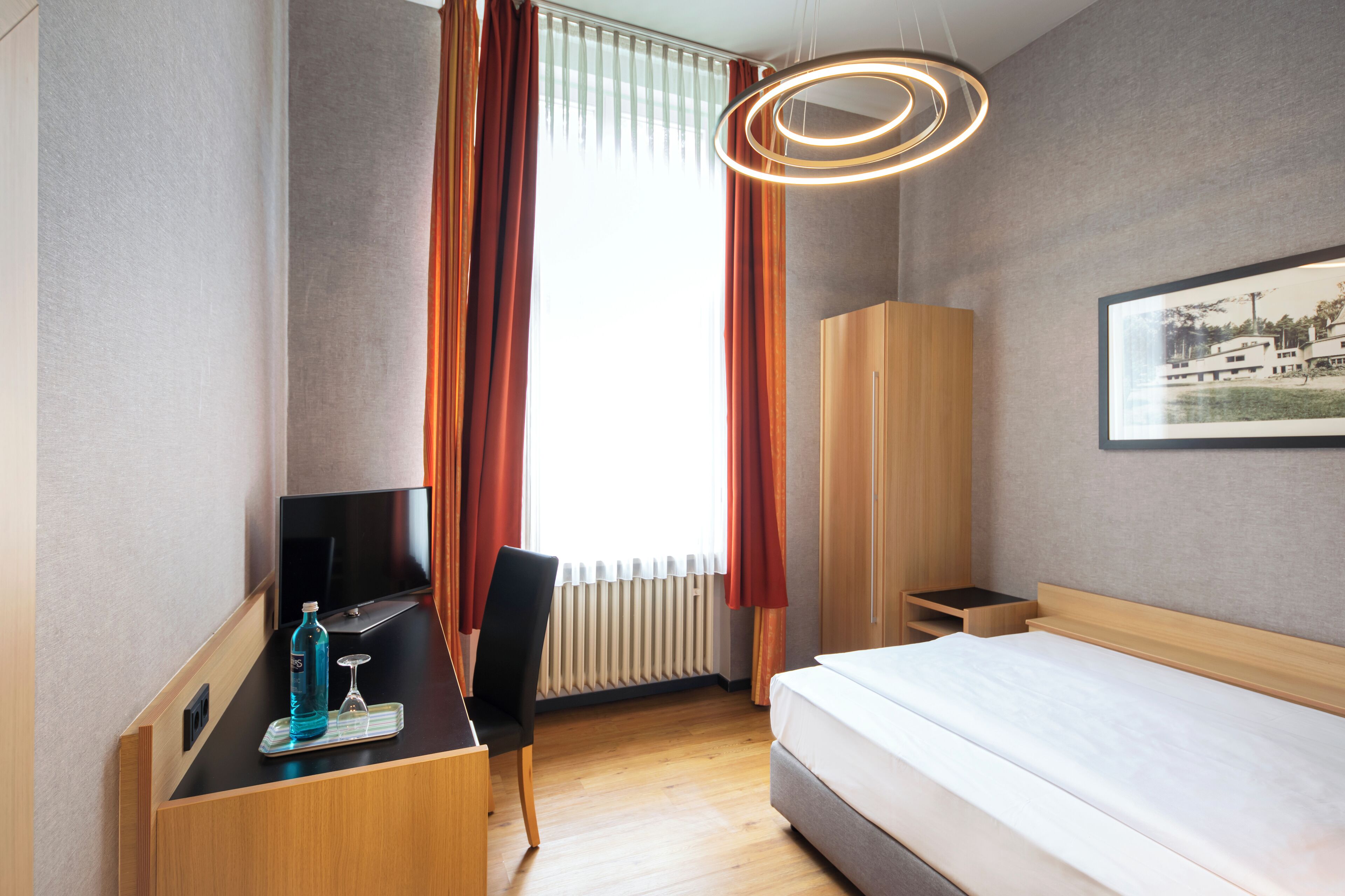 Une chambre d’hôtel avec un lit, un bureau avec une télévision, une chaise, un radiateur et un tableau au mur.