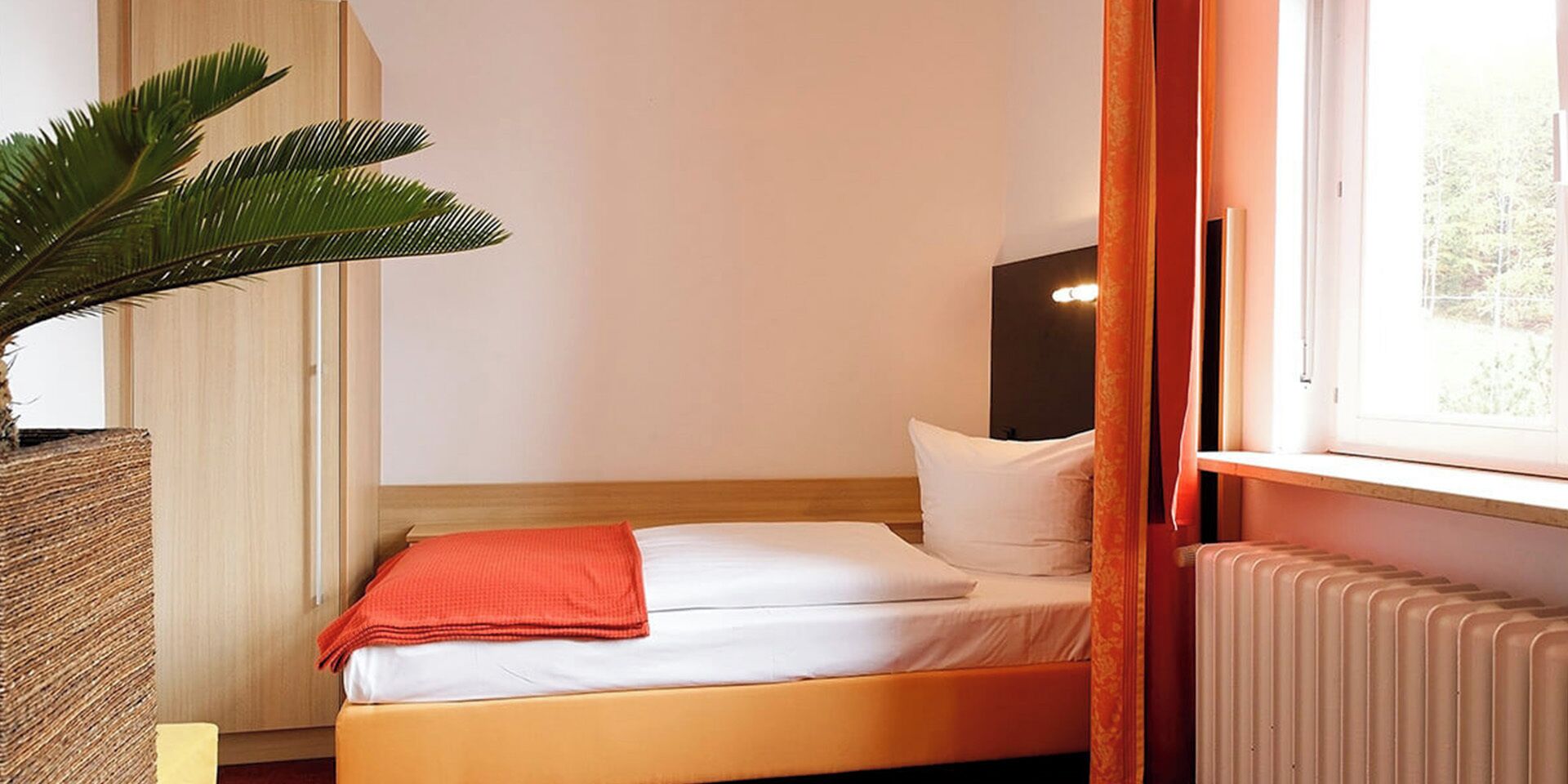 A single bed with white bedding and an orange throw, a radiator, a potted plant, and a window with curtains.