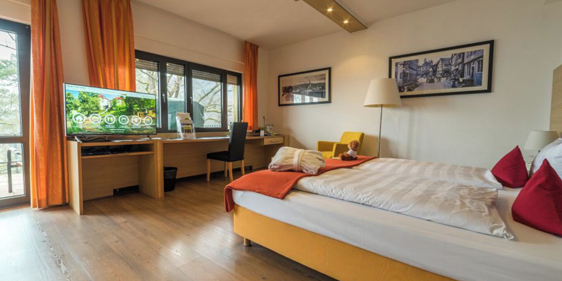 Habitación de hotel moderna con una cama grande, un escritorio con computadora, un televisor y una ventana con cortinas.