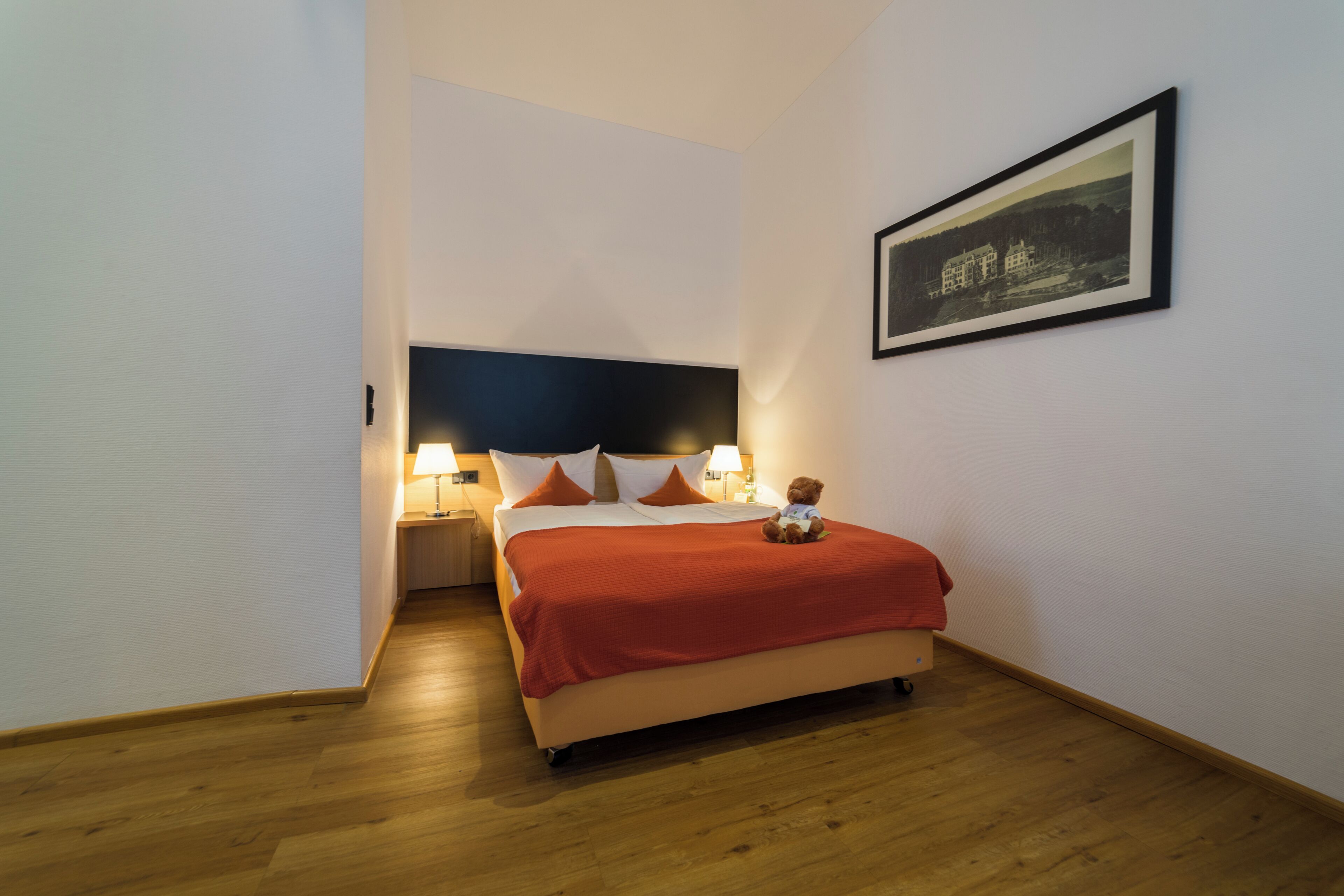 A hotel room with a bed, a nightstand, a lamp, a teddy bear, and a framed picture on the wall.