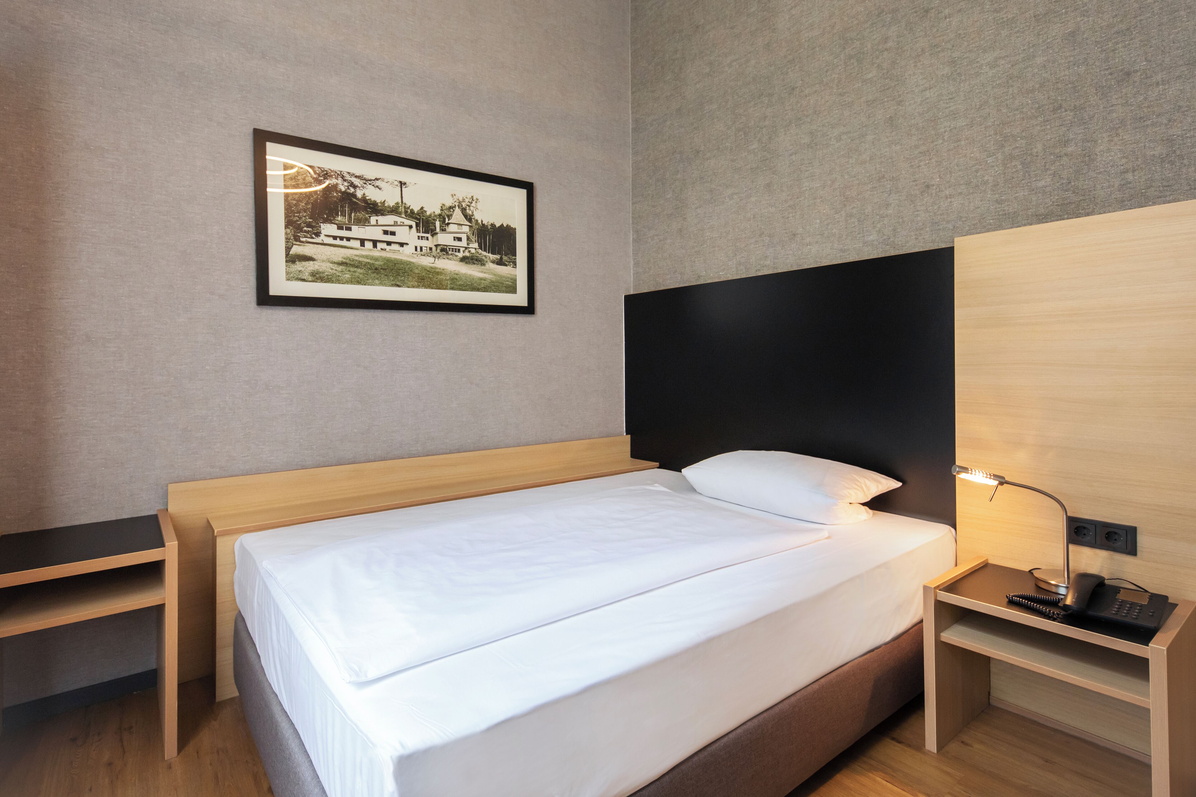 A neatly made bed with a white mattress and pillow, a bedside table with a lamp, and a framed picture on the wall.