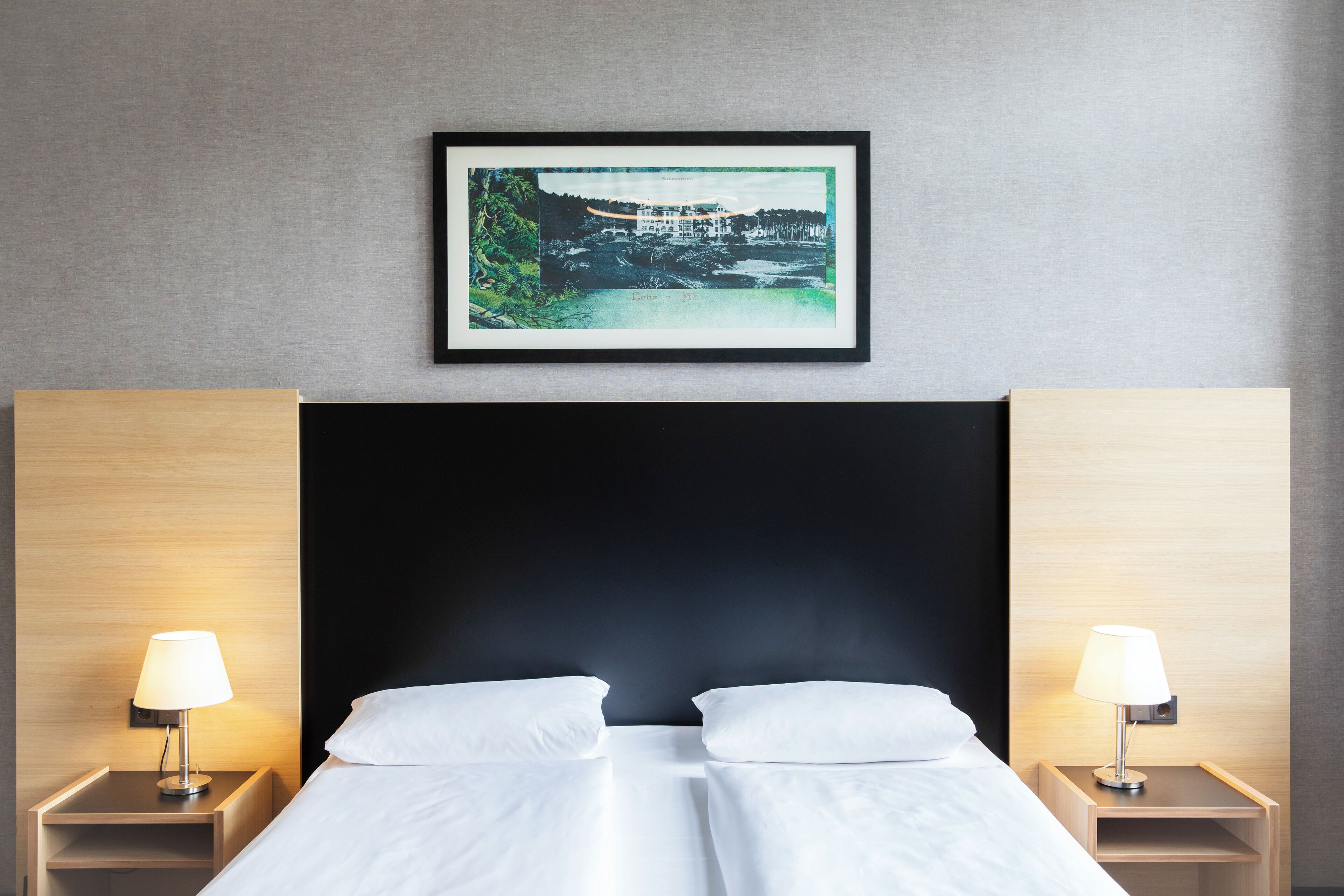 A hotel room with a large bed, two bedside tables with lamps, a framed picture on the wall, and a dark headboard.
