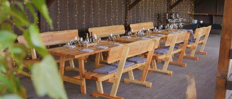 Un coin repas rustique en bois, avec des bancs et des tables, des guirlandes lumineuses et des verres à vin.