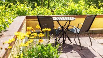 Un patio al aire libre con una mesa redonda y dos sillas, rodeado de follaje y flores amarillas.