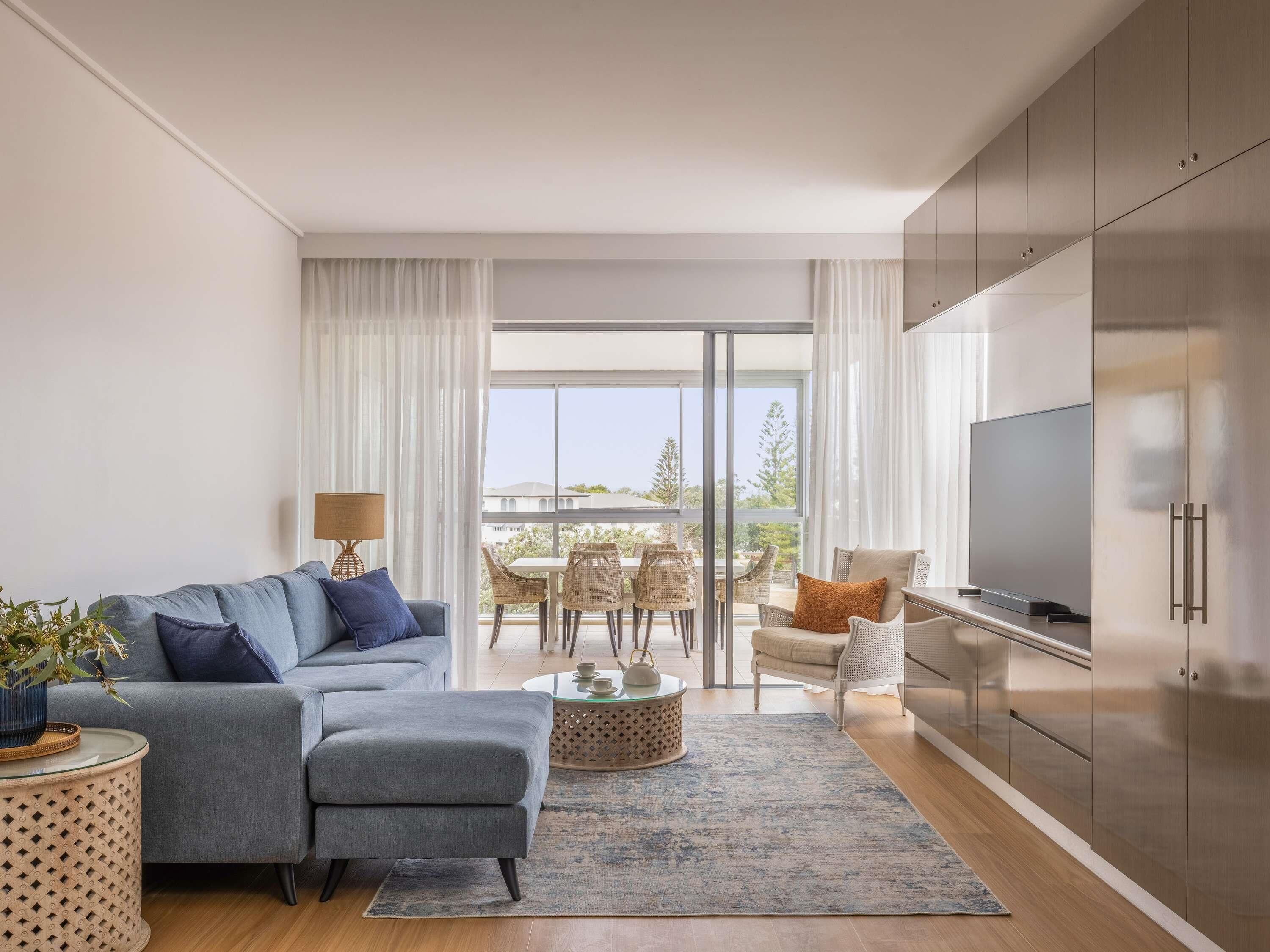 A modern living room with a sofa, a coffee table, and a TV. There is a balcony with outdoor furniture.