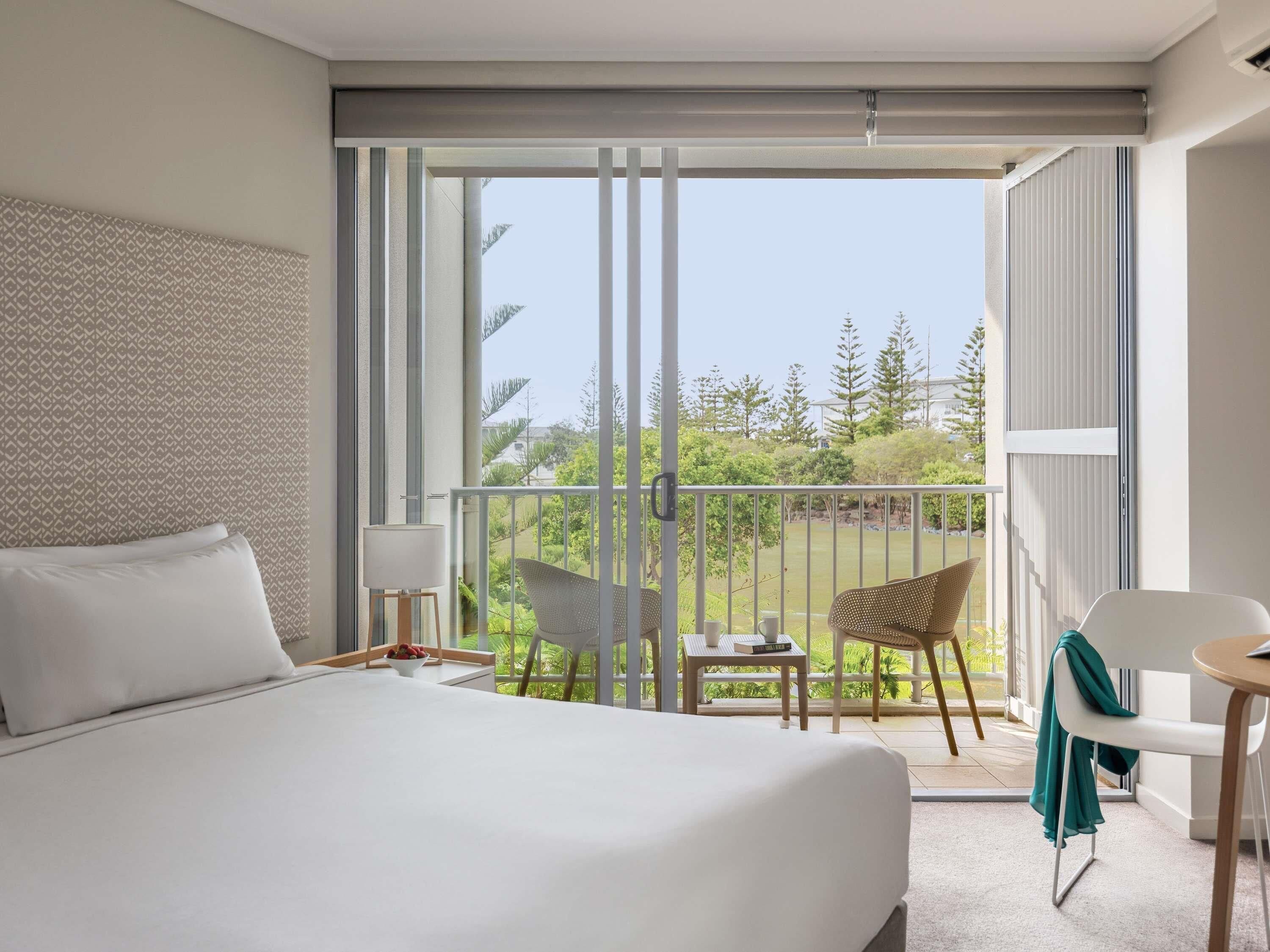 A hotel room with a large bed, a balcony with a chair and table, and a view of greenery.