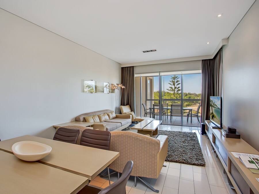 A modern living room with a sofa, dining table, and chairs. There is a large window with curtains, a flat-screen TV, and a view of greenery outside.