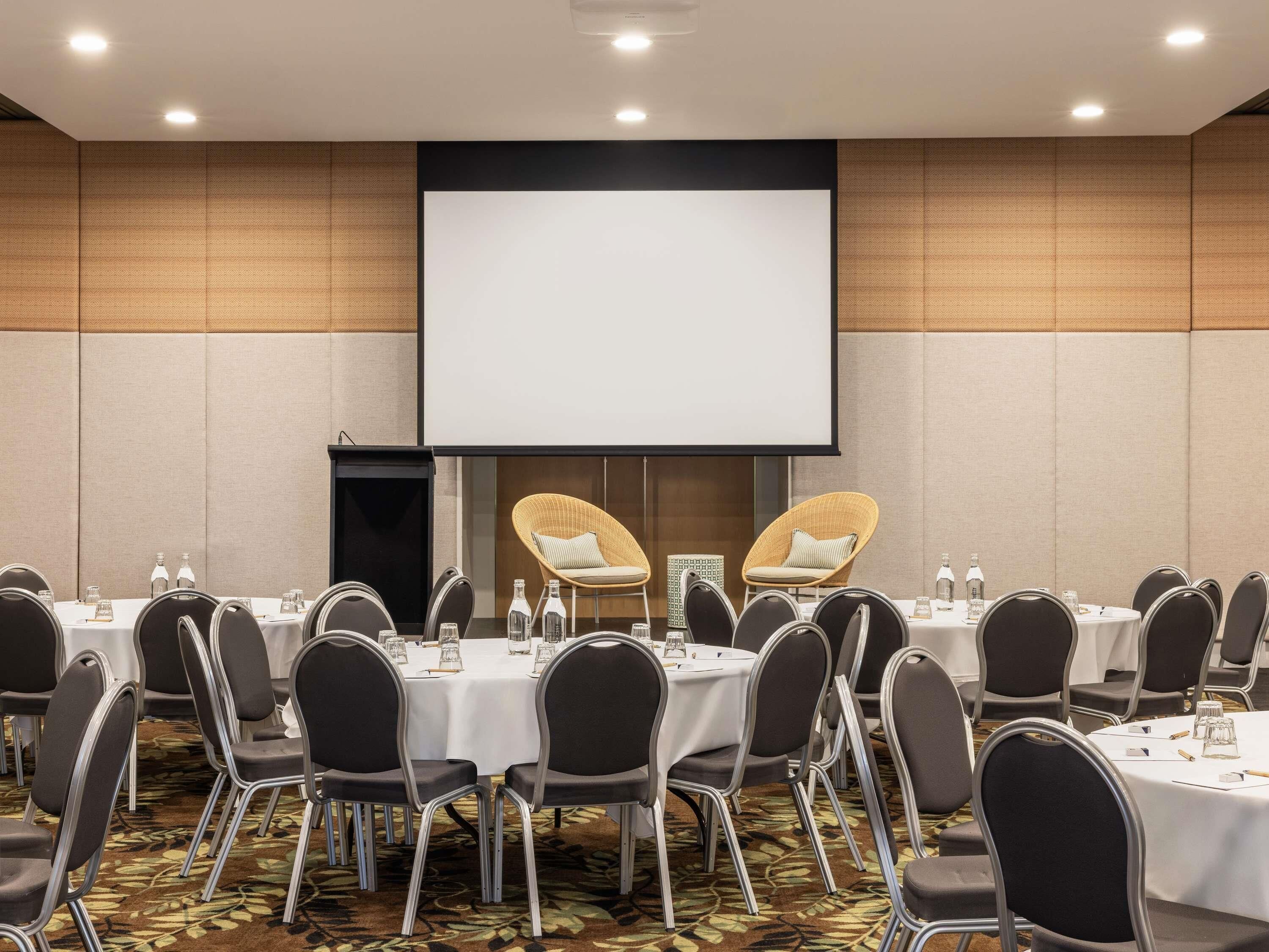 Un salón de conferencias con mesas redondas, sillas negras, una pantalla grande y un atril.