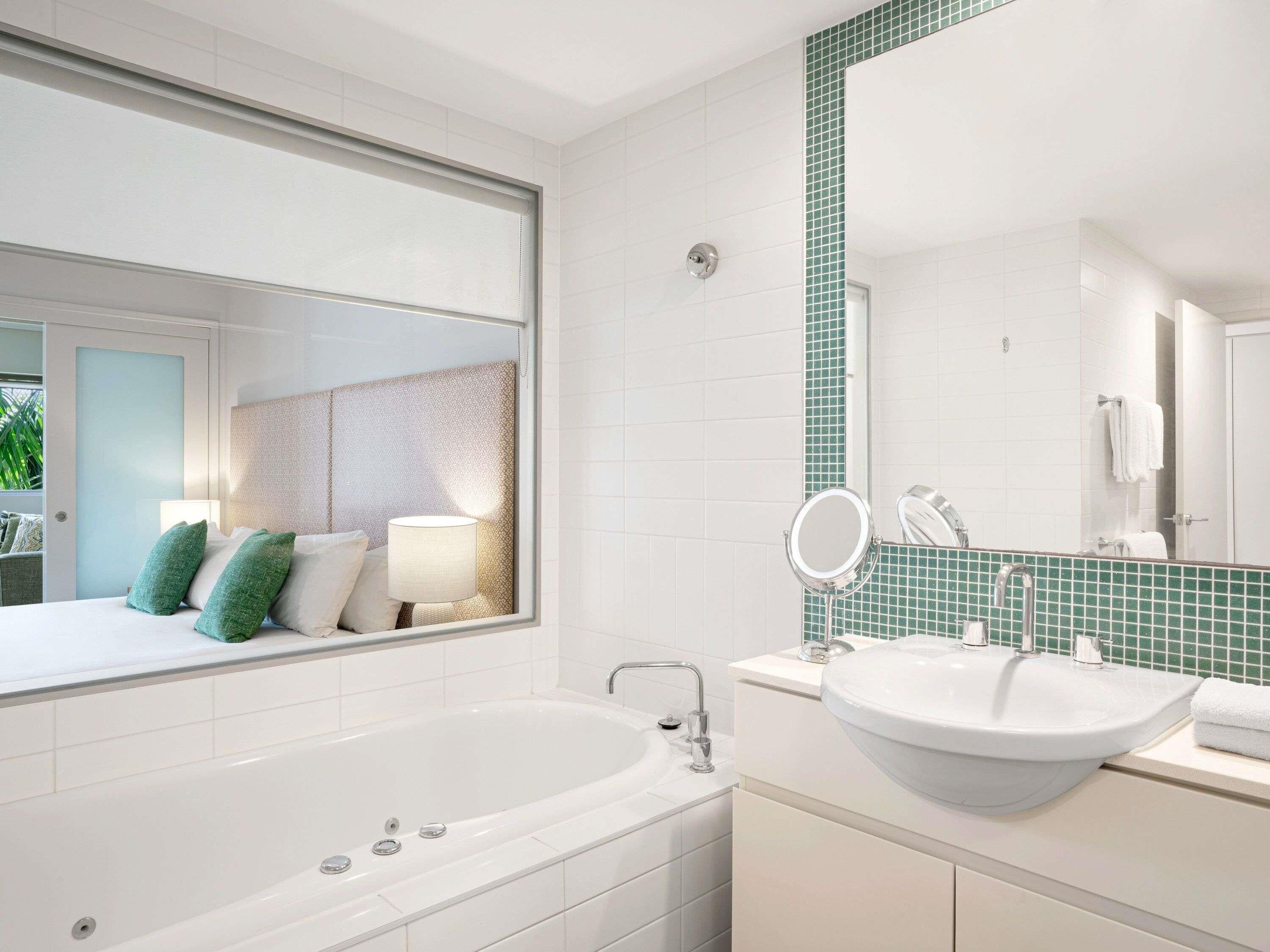 A modern bathroom with a large bathtub, a sink with a mirror, and a window overlooking a bedroom.