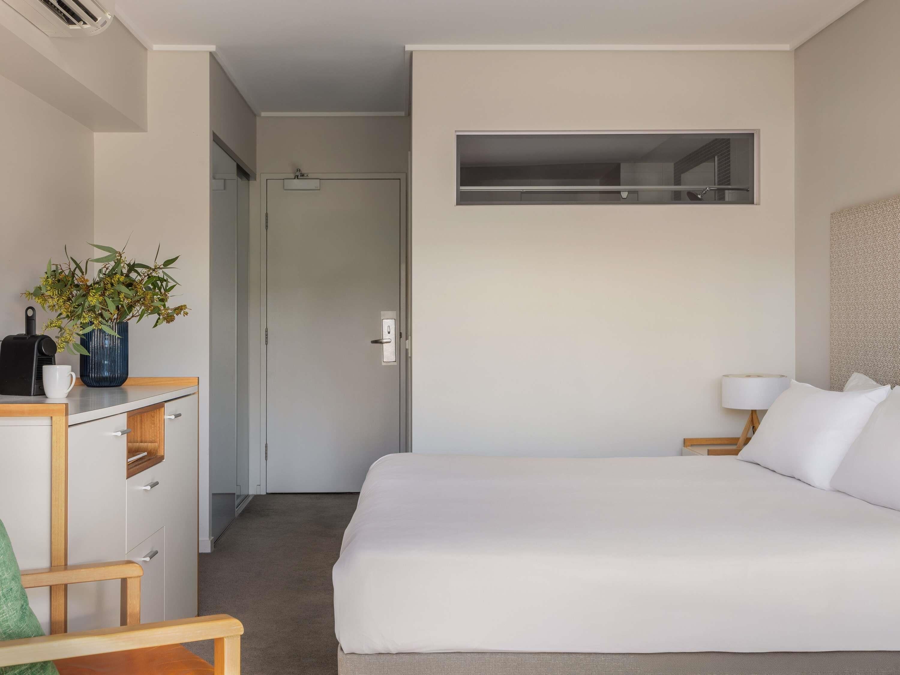 A hotel room with a bed, a desk with a plant, a lamp, and a view of the hallway.