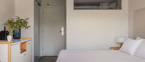A hotel room with a bed, a desk with a plant, a lamp, and a view of the hallway.