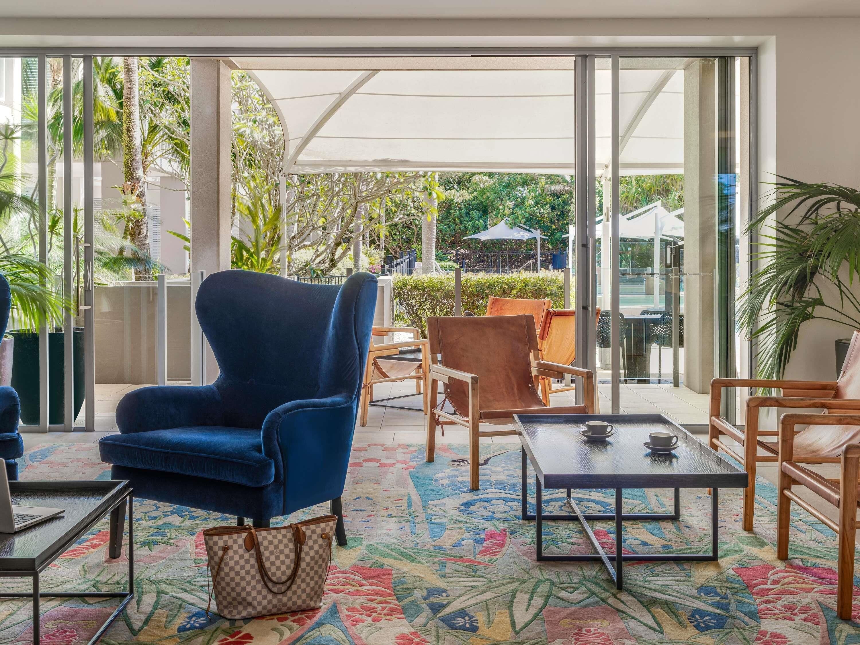 Een moderne zithoek met een blauwe fauteuil, houten stoelen, een kamerbreed tapijt en een glazen deur die leidt naar een buitenste tuin.
