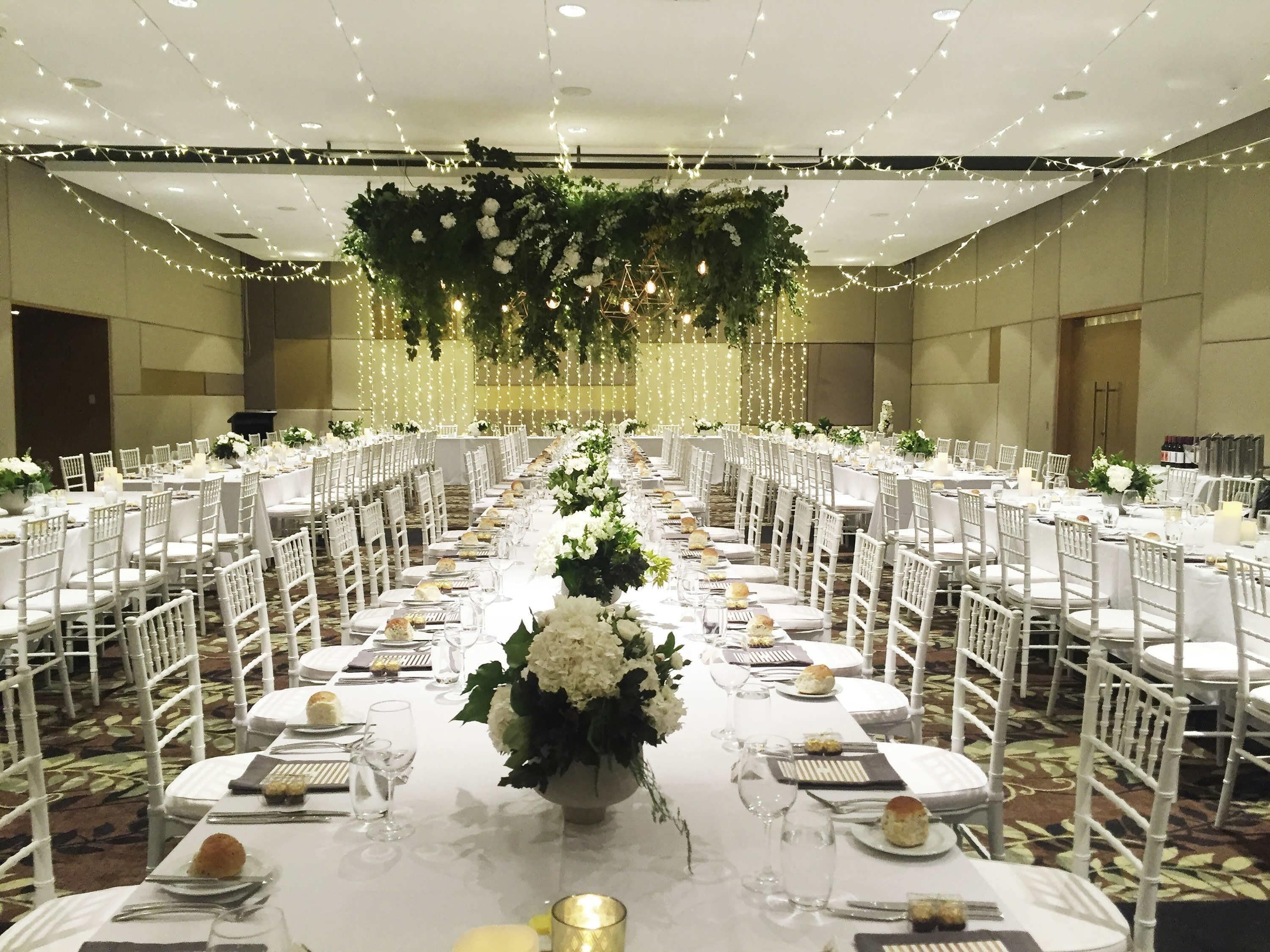 A banquet hall with white tables and chairs, floral centerpieces, and string lights.