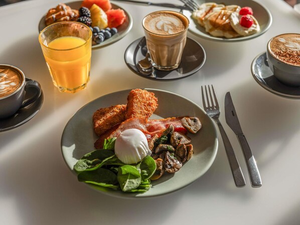 Een ontbijttafel met croissants, fruit, koffie, en een bord met spek, champignons, spinazie en een gepocheerd ei.