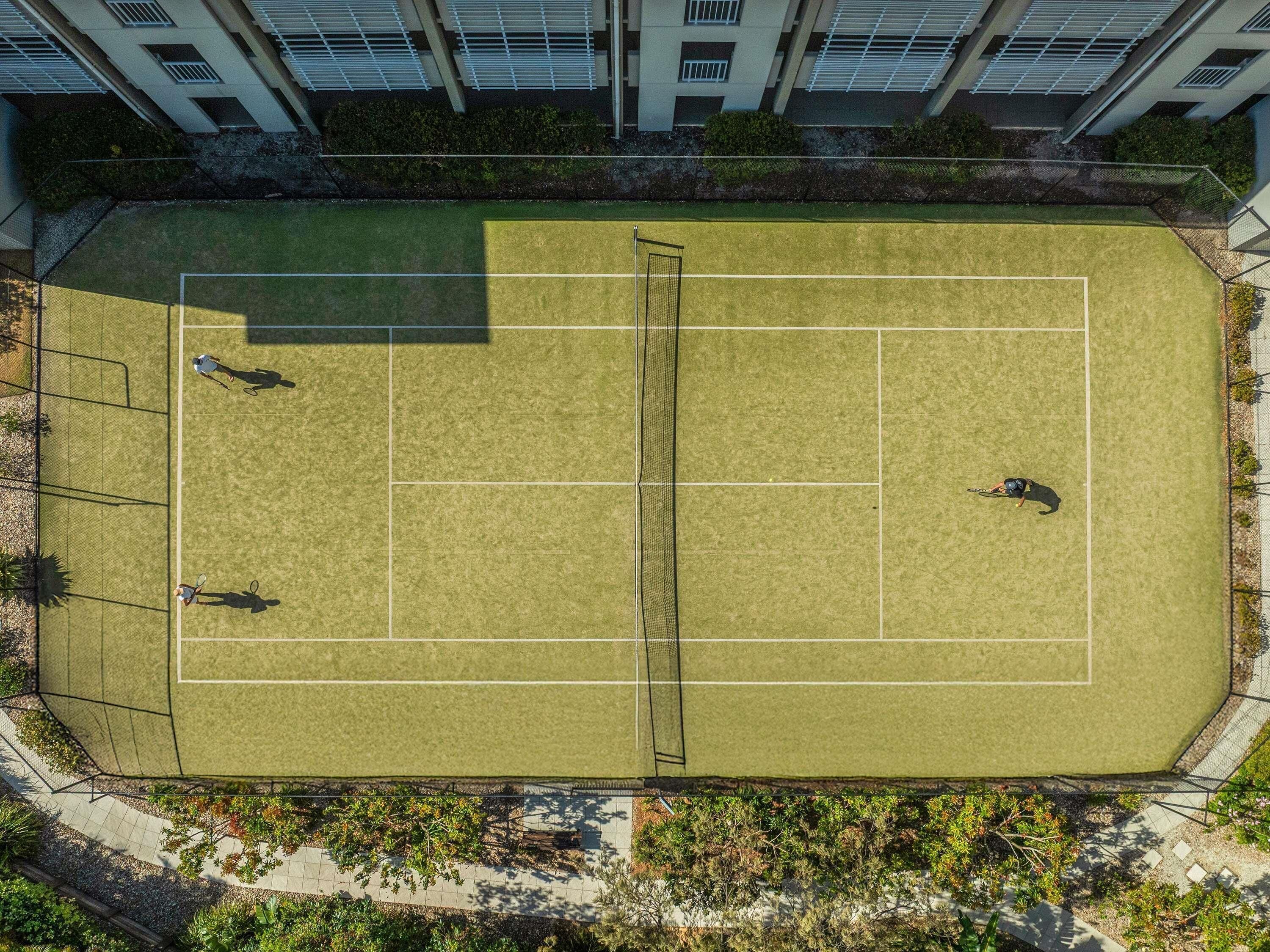 An aerial view of a tennis court surrounded by a fence, with buildings in the background.