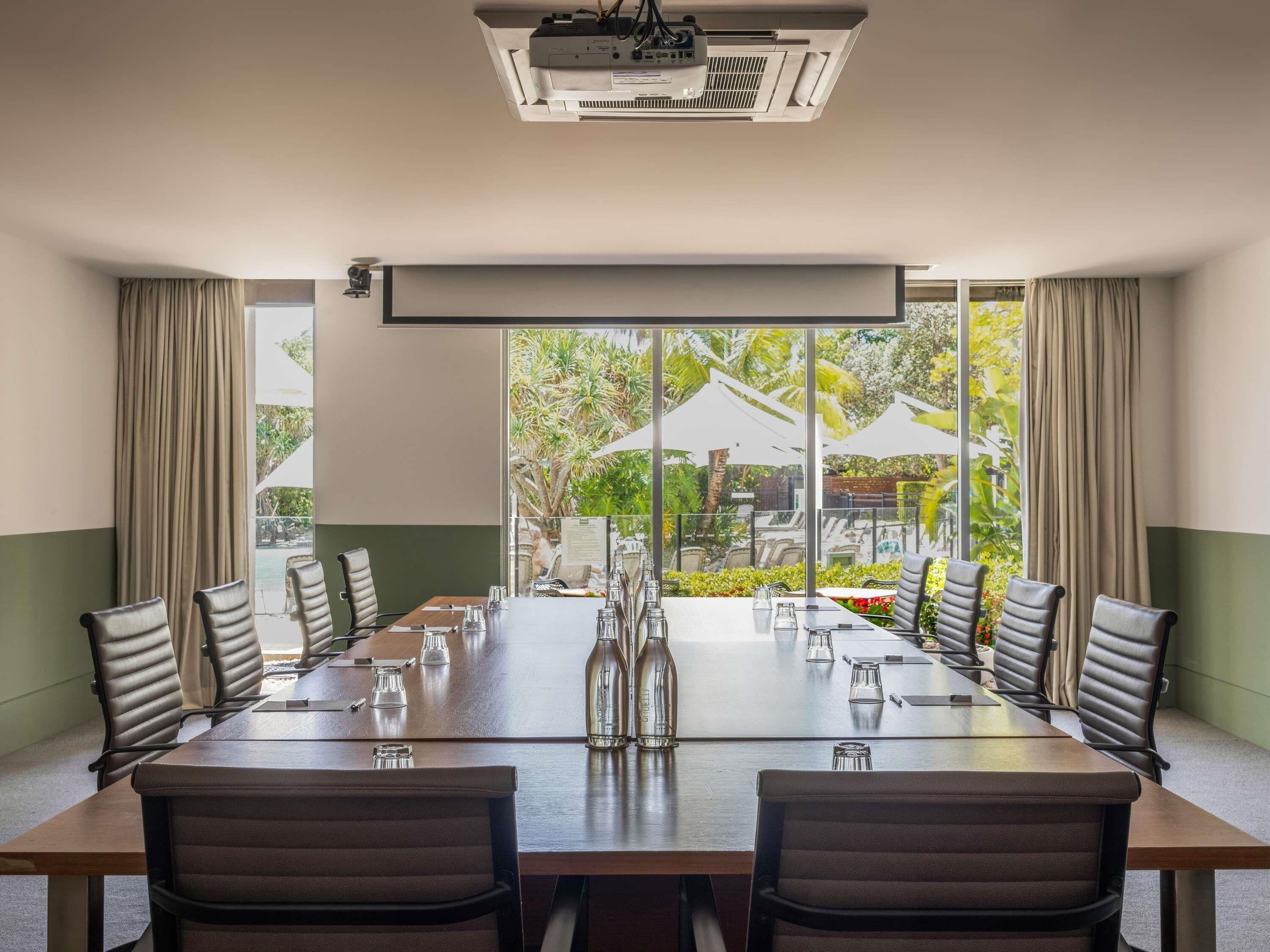 A conference room with a long table, chairs, a projector, and a view of an outdoor garden.