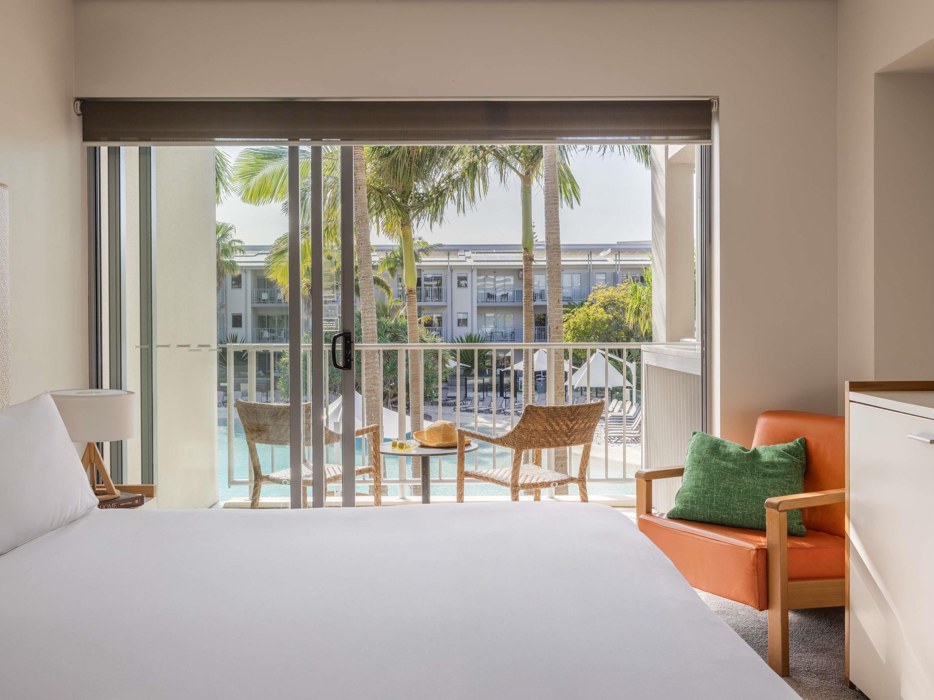 Een hotelkamer met een groot bed, een balkon met uitzicht op het zwembad en twee rieten stoelen.