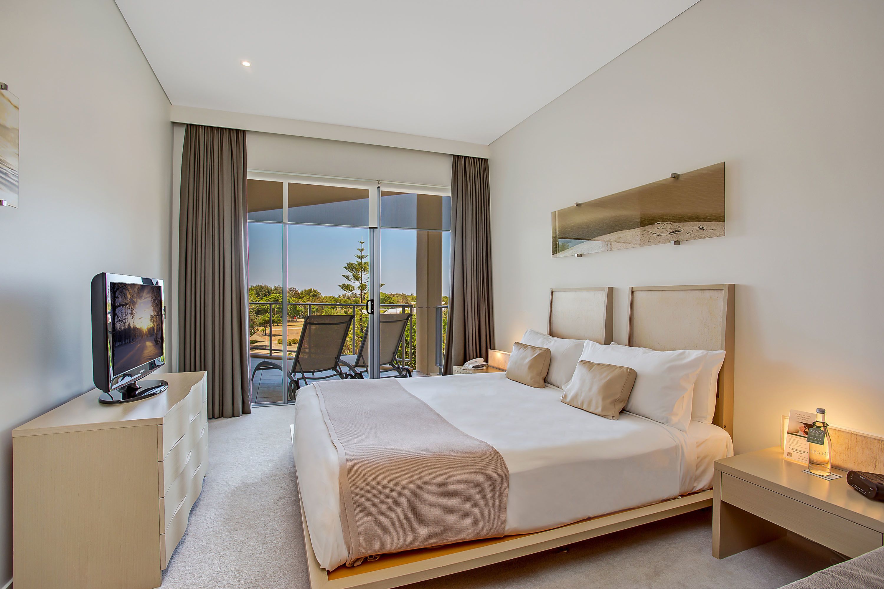 Une chambre d’hôtel moderne avec un grand lit, un téléviseur posé sur une commode, une table de chevet avec une lampe, et une vue sur l’extérieur grâce à une porte coulissante.