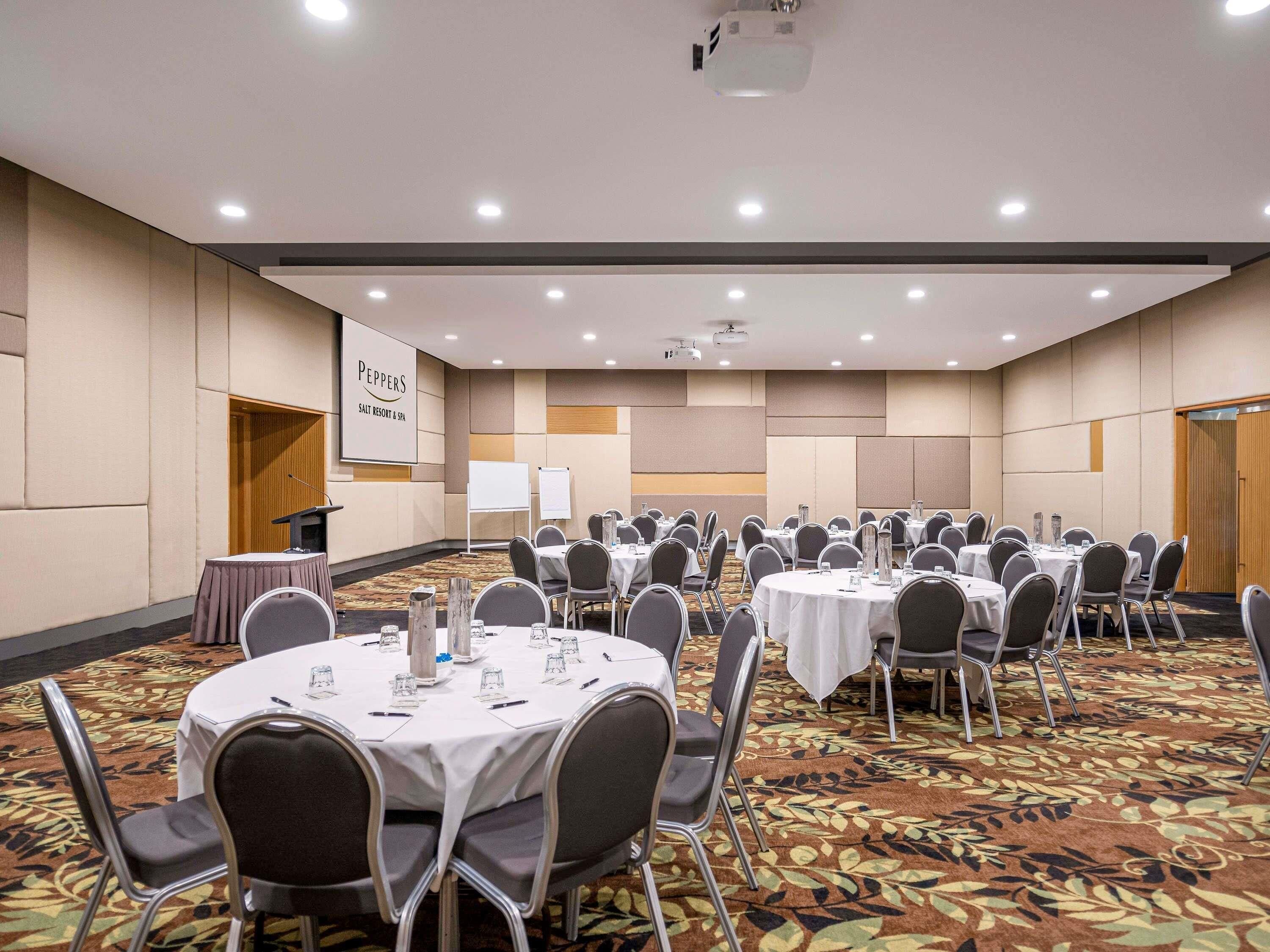 A conference room with multiple round tables, chairs, a podium, and a presentation screen.