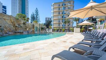 A pool area with lounge chairs and umbrellas, surrounded by buildings and a rocky landscape.