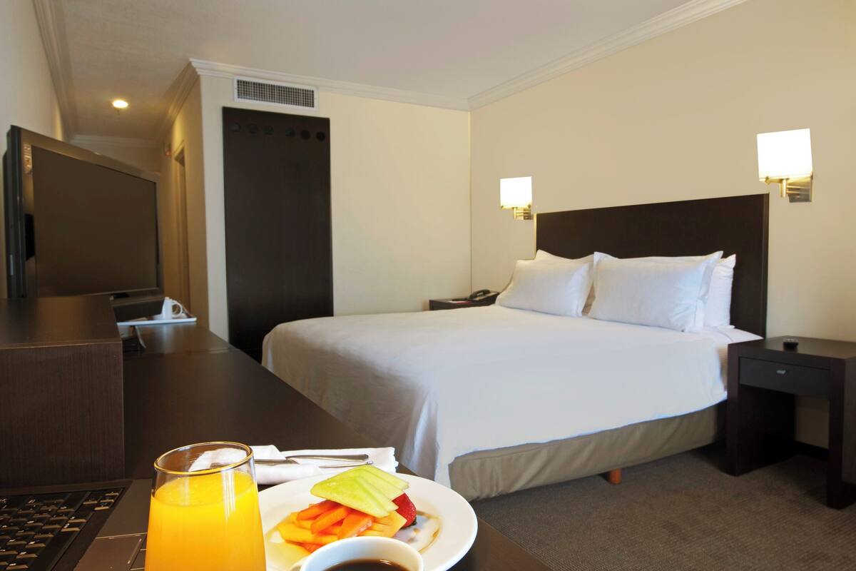 A hotel room with a bed, bedside table, and a desk with a laptop and breakfast plate.
