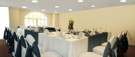 Un salón de conferencias con mesas redondas, sillas blancas con cintas azules y un centro de mesa floral.