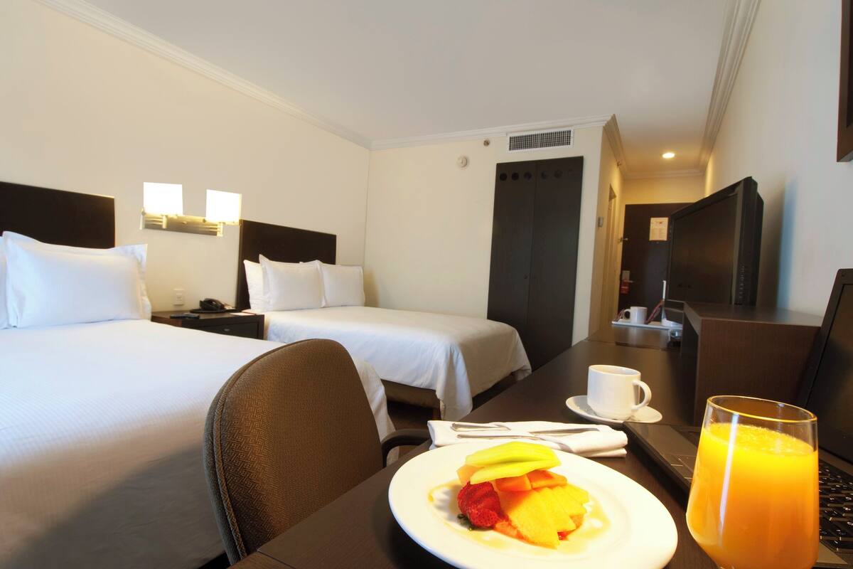 A hotel room with two beds, a dining table with breakfast, a laptop, and a glass of orange juice.