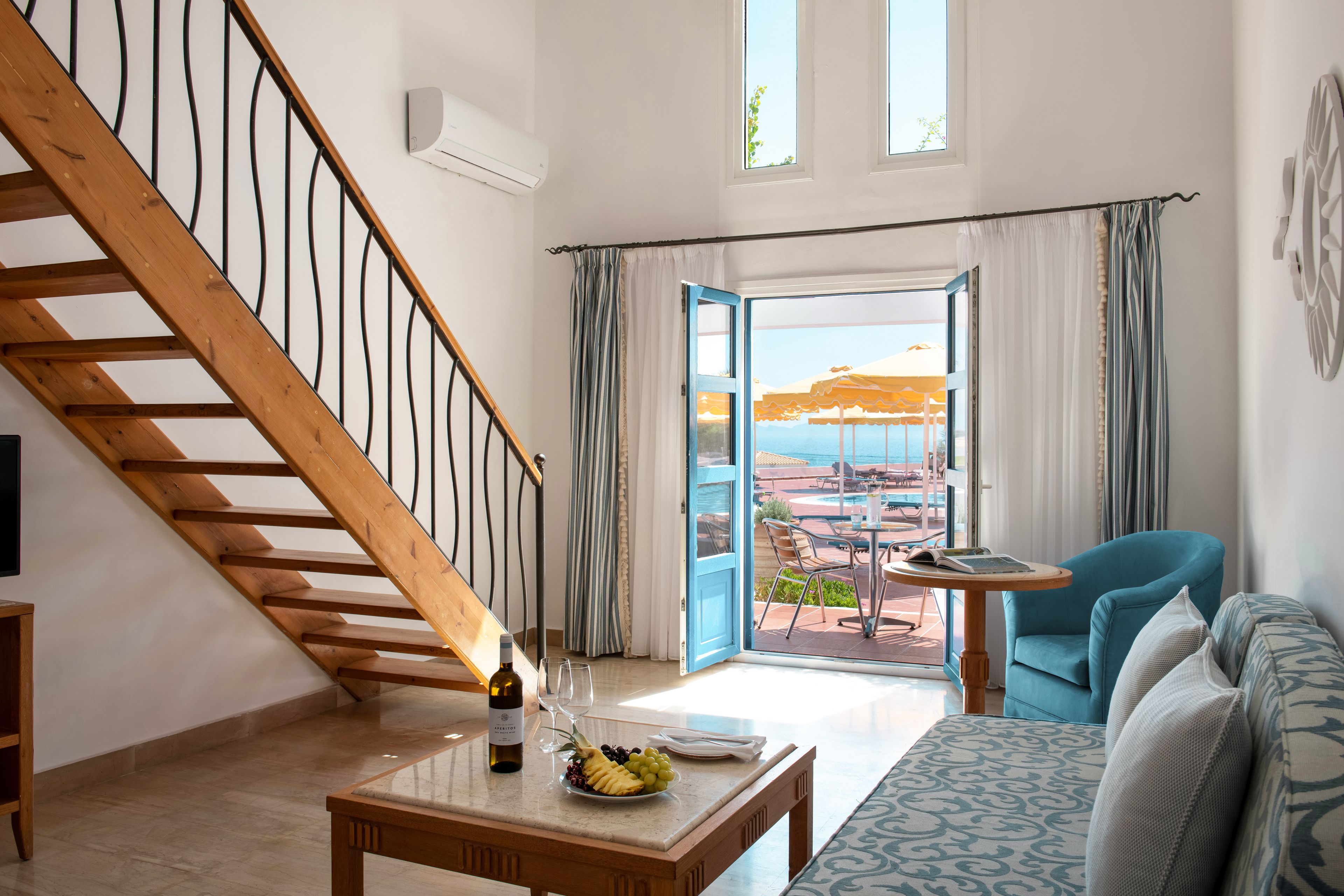 Una habitación con una escalera de madera, una mesa de comedor con una botella y vasos, un sofá y vistas a una piscina y sombrillas a través de una puerta abierta.