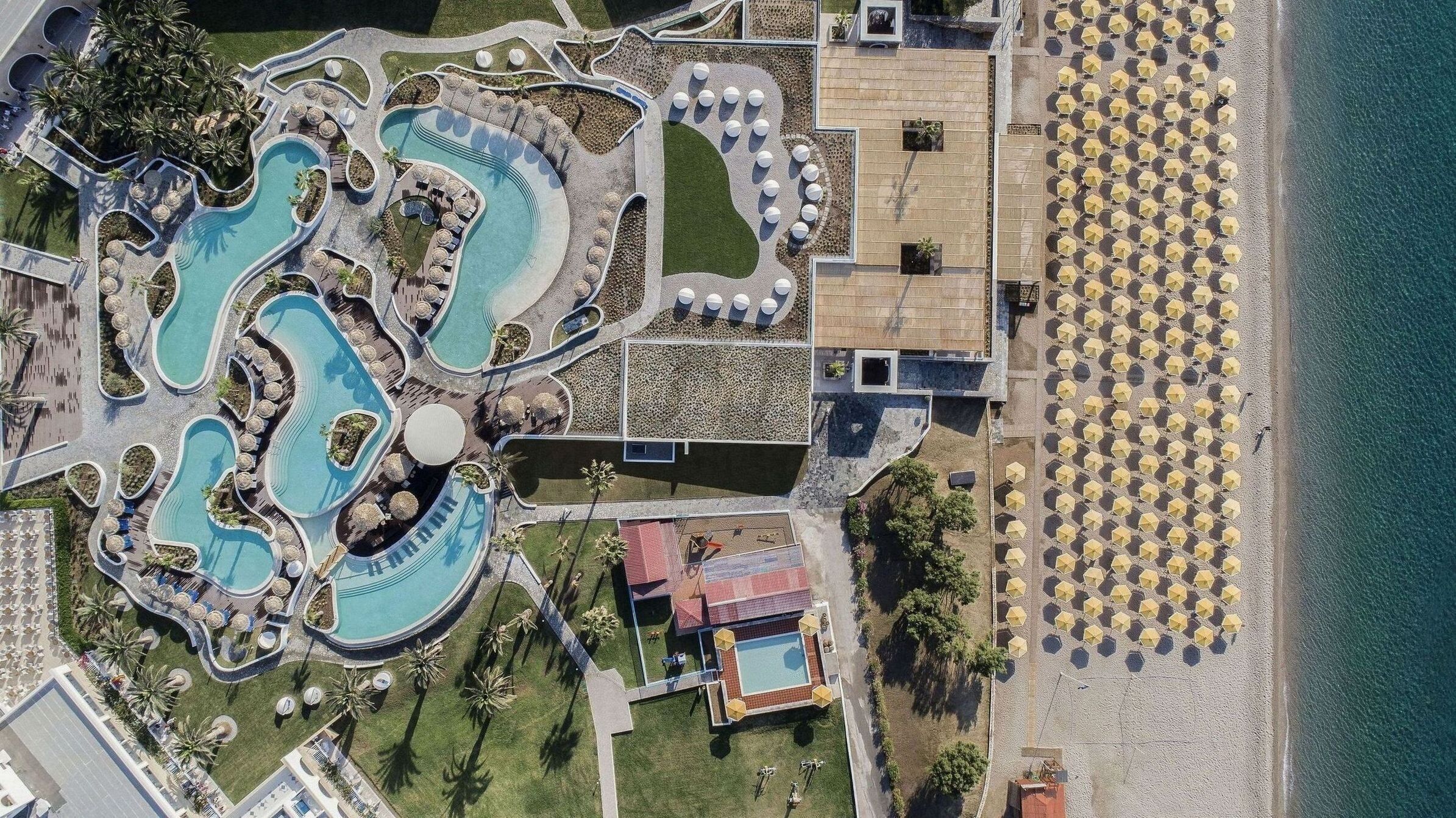 Luchtbeeld van een resort met meerdere zwembaden, een strand met parasols en een gebouw met een rood dak.