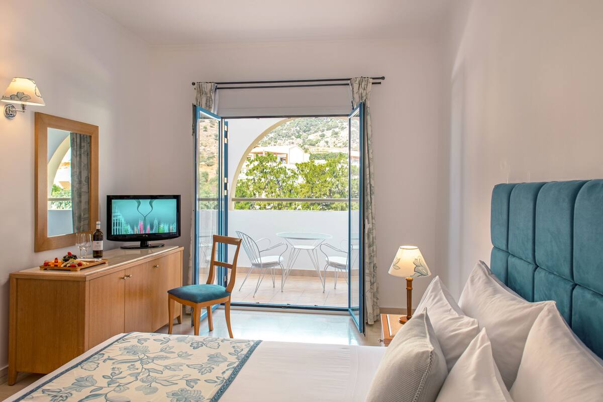 Ein Hotelzimmer mit Bett, Fernseher, einem Holzschrank, einem Sessel, einem Tisch mit Stühlen und einem Balkon mit Blick auf Grünflächen und Gebäude.