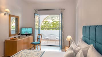 Une chambre d’hôtel comprenant un lit, une télévision, une commode en bois, une chaise, une table avec des chaises et un balcon donnant sur un paysage verdoyant et des bâtiments.