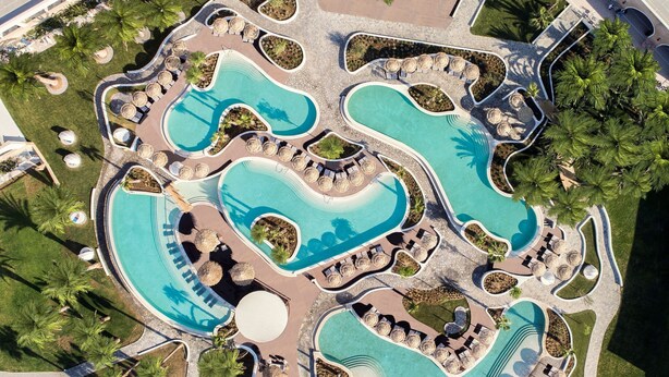 An aerial view of a resort with multiple interconnected swimming pools, lounge areas, and palm trees. - Mitsis Norida (Kos)
