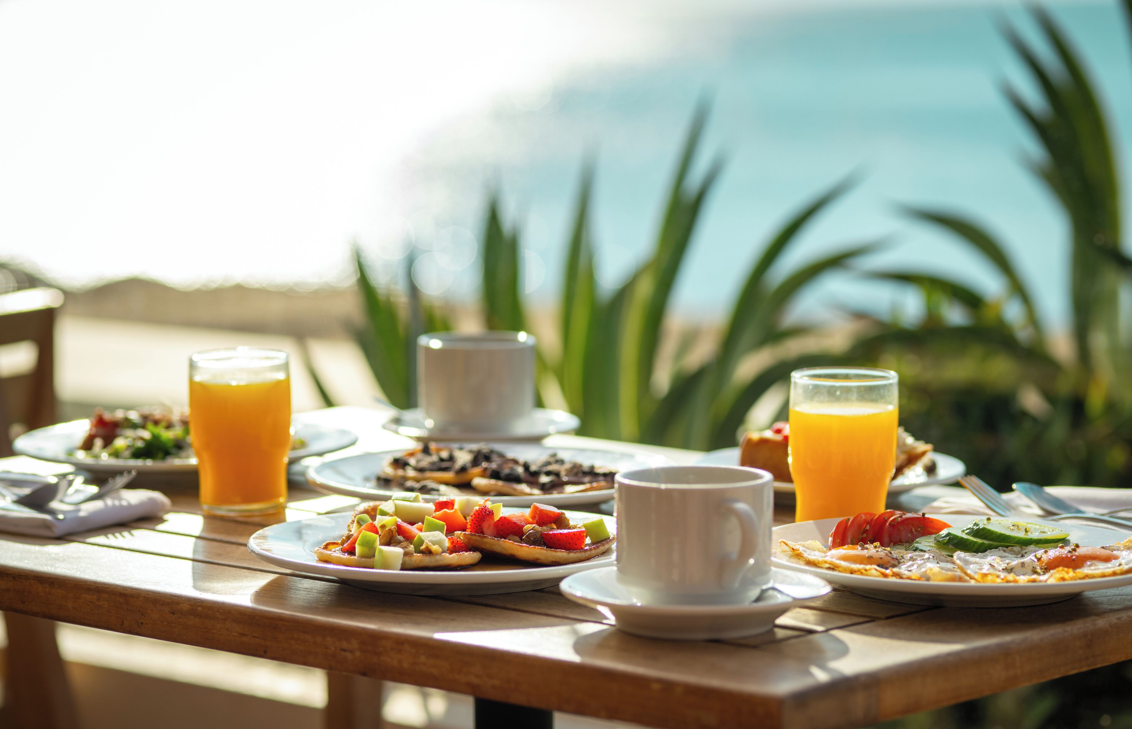 Ett frukostbord med tallrikar fyllda med mat, glas med apelsinjuice och en kopp, placerat utomhus med utsikt över havet.