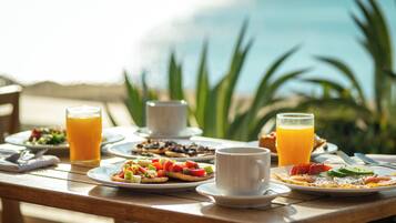 A breakfast table with plates of food, glasses of orange juice, and a cup, set outdoors with a view of the ocean.