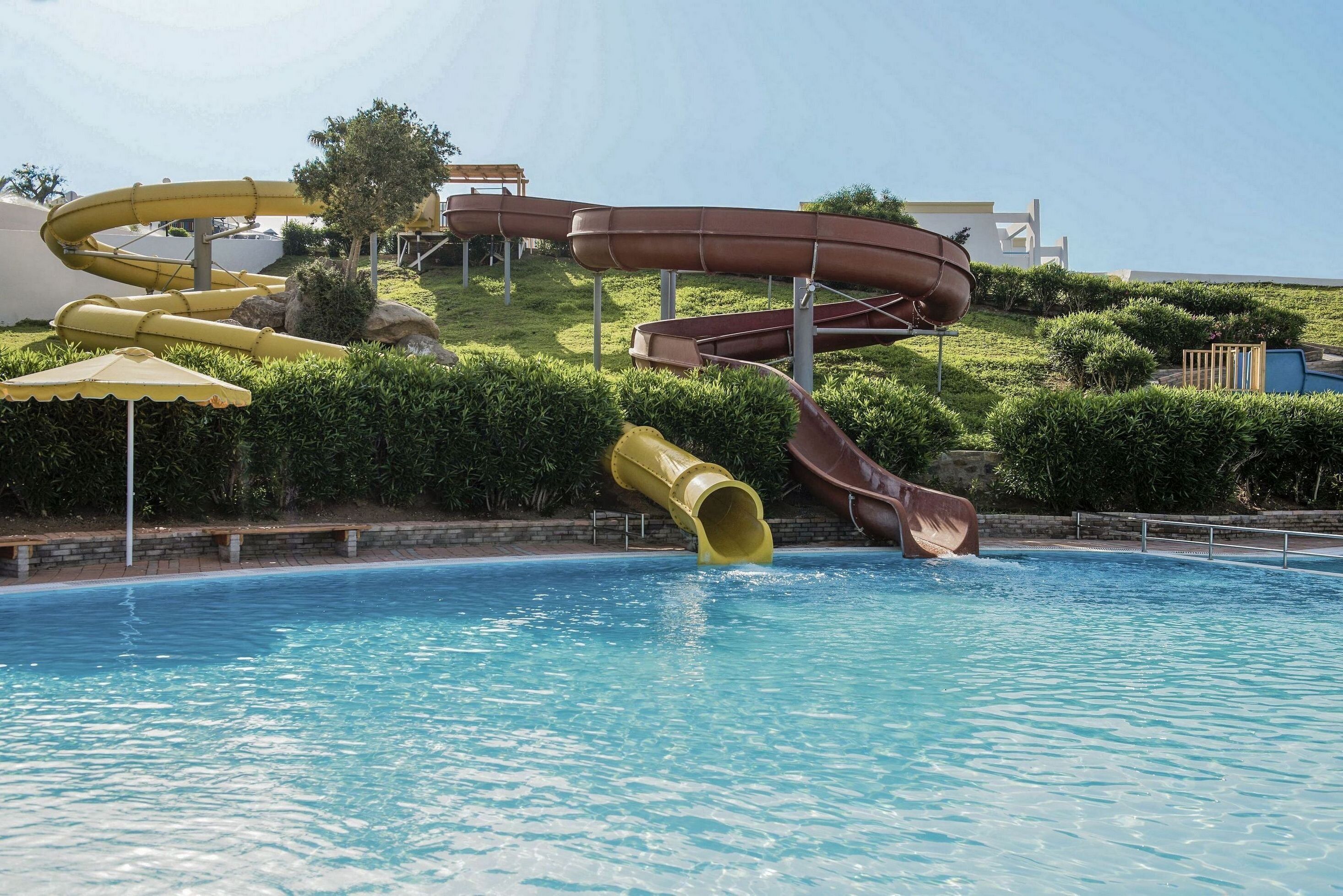 Una piscina con diversi scivoli, tra cui uno giallo e uno marrone, e una zona a bordo piscina con una panchina e un ombrellone.