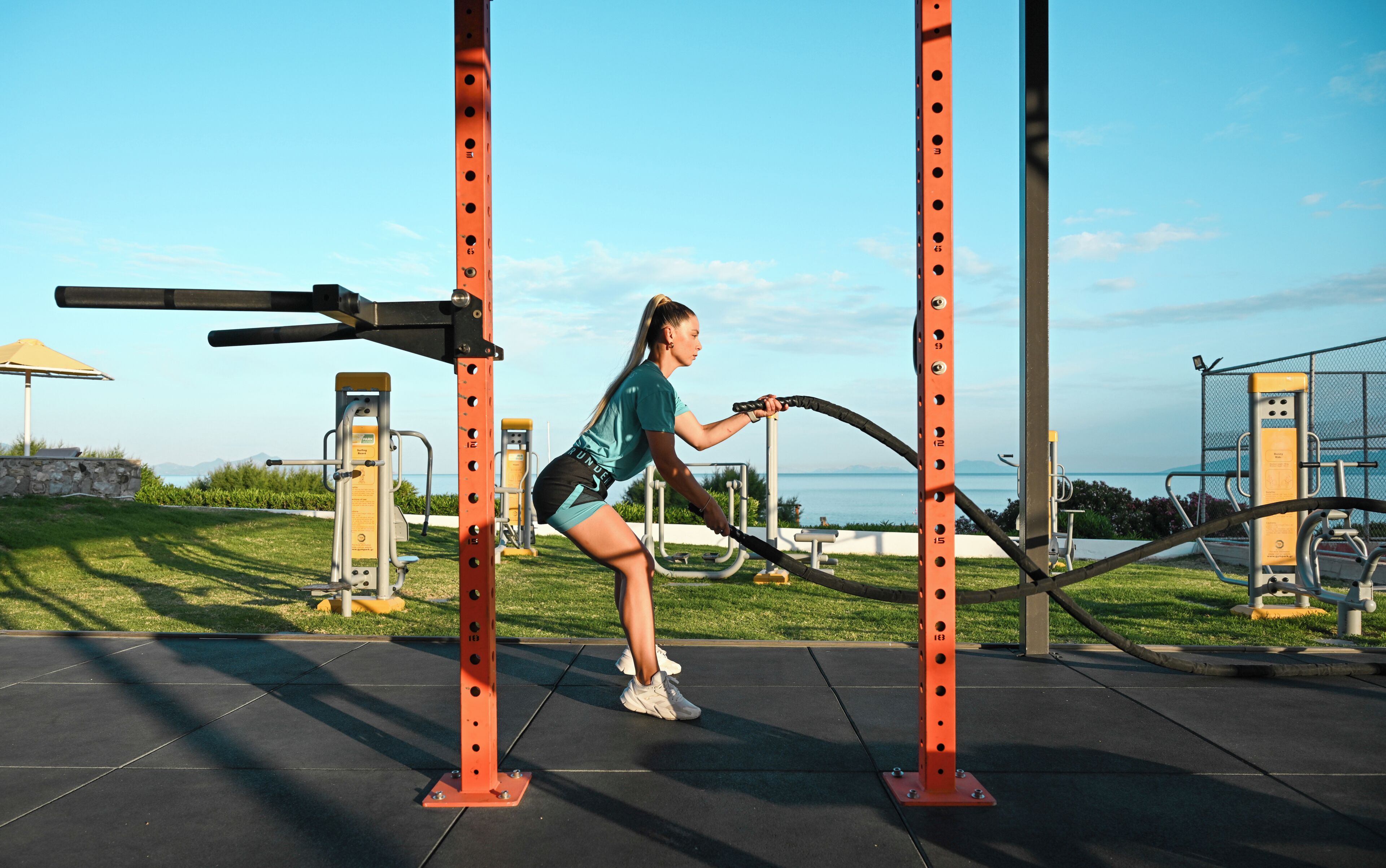 Una persona ejercitándose con una máquina de cuerdas de batalla en un gimnasio al aire libre, con equipo de ejercicio y vistas al mar.