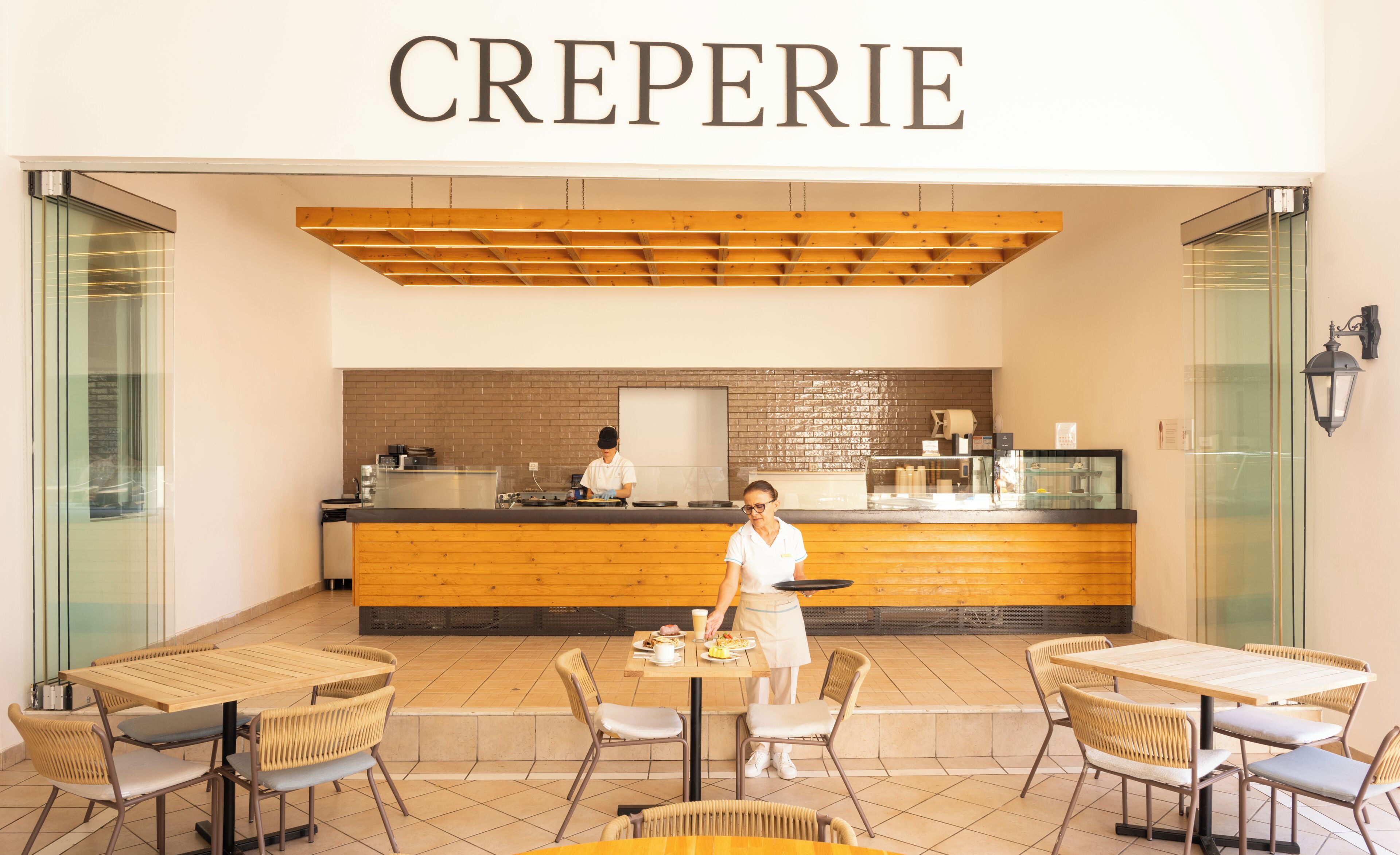 A cafe named 'CREPERIE' with a wooden counter, a person serving, and outdoor seating.