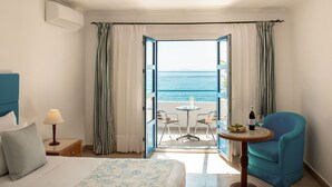 A hotel room with a bed, a table with a bottle and glasses, a chair, and a view of the sea through an open door. - Mitsis Norida (Kos)
