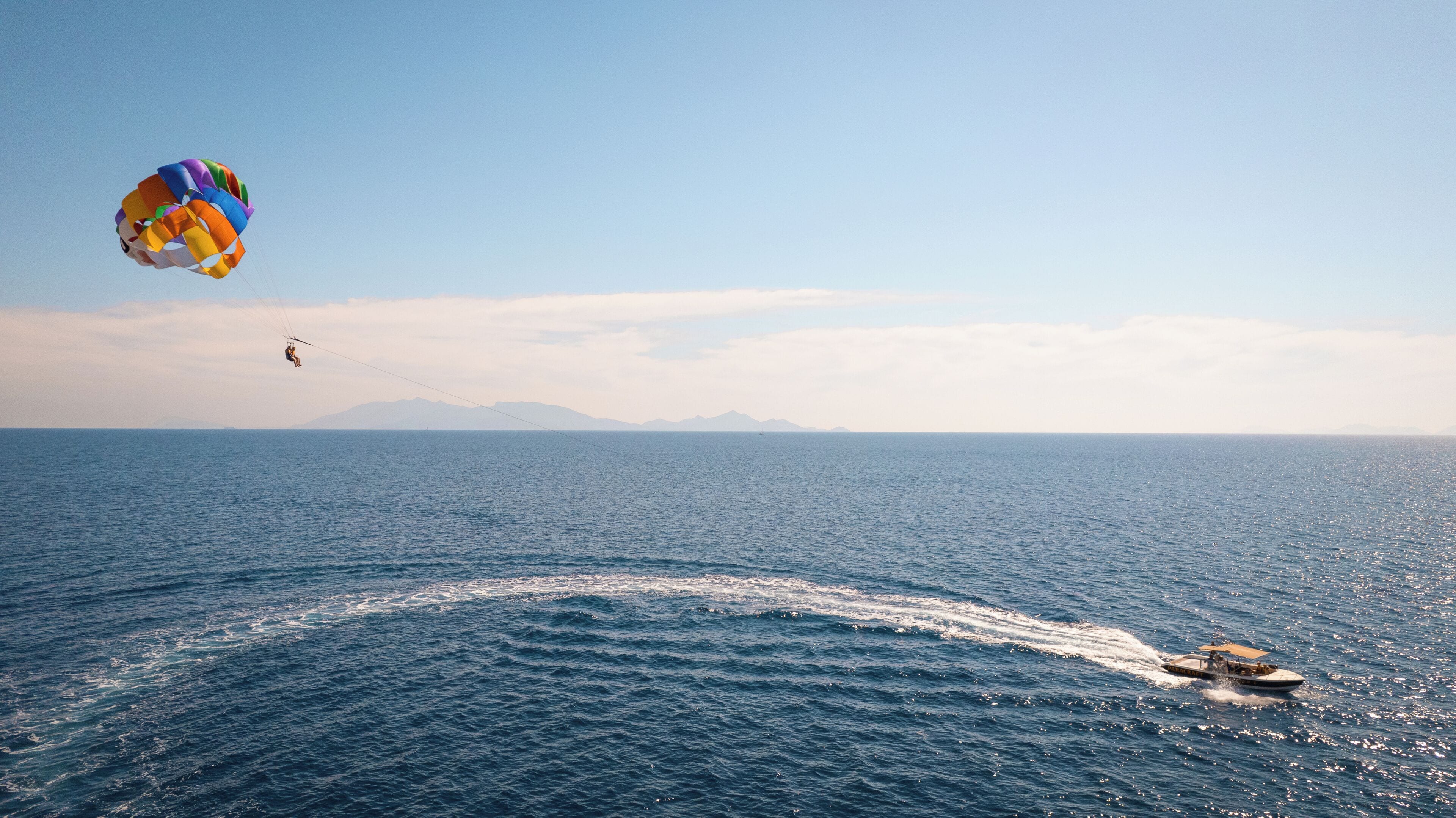 Vista despejada del océano con un barco dejando una estela, un paracaídas en el aire y montañas distantes bajo un cielo claro.