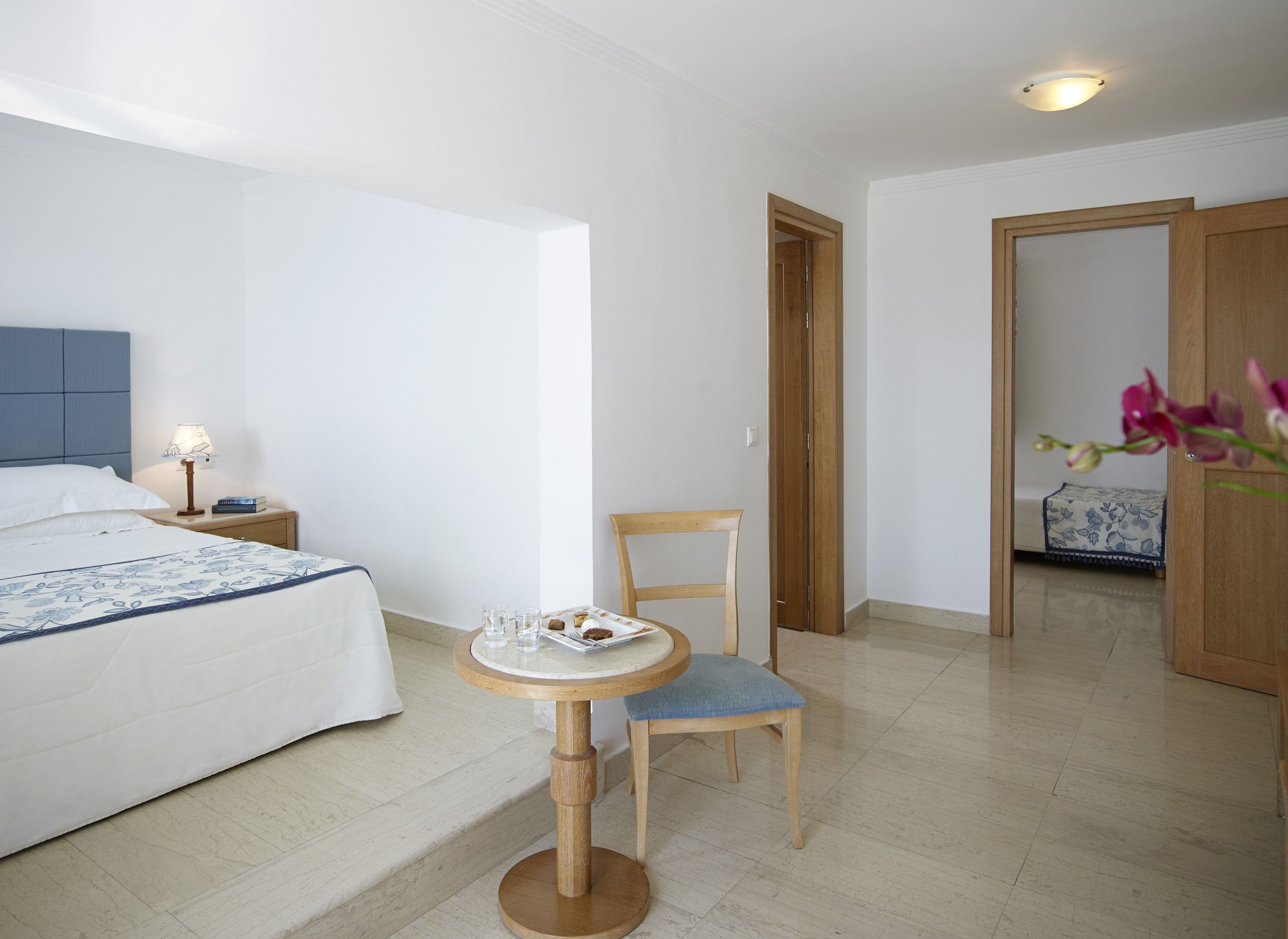 A hotel room with a bed, bedside table with a lamp, a small round table with a chair, and a wooden door leading to another room.