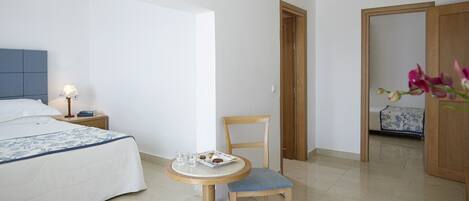 A hotel room with a bed, bedside table with a lamp, a small round table with a chair, and a wooden door leading to another room.