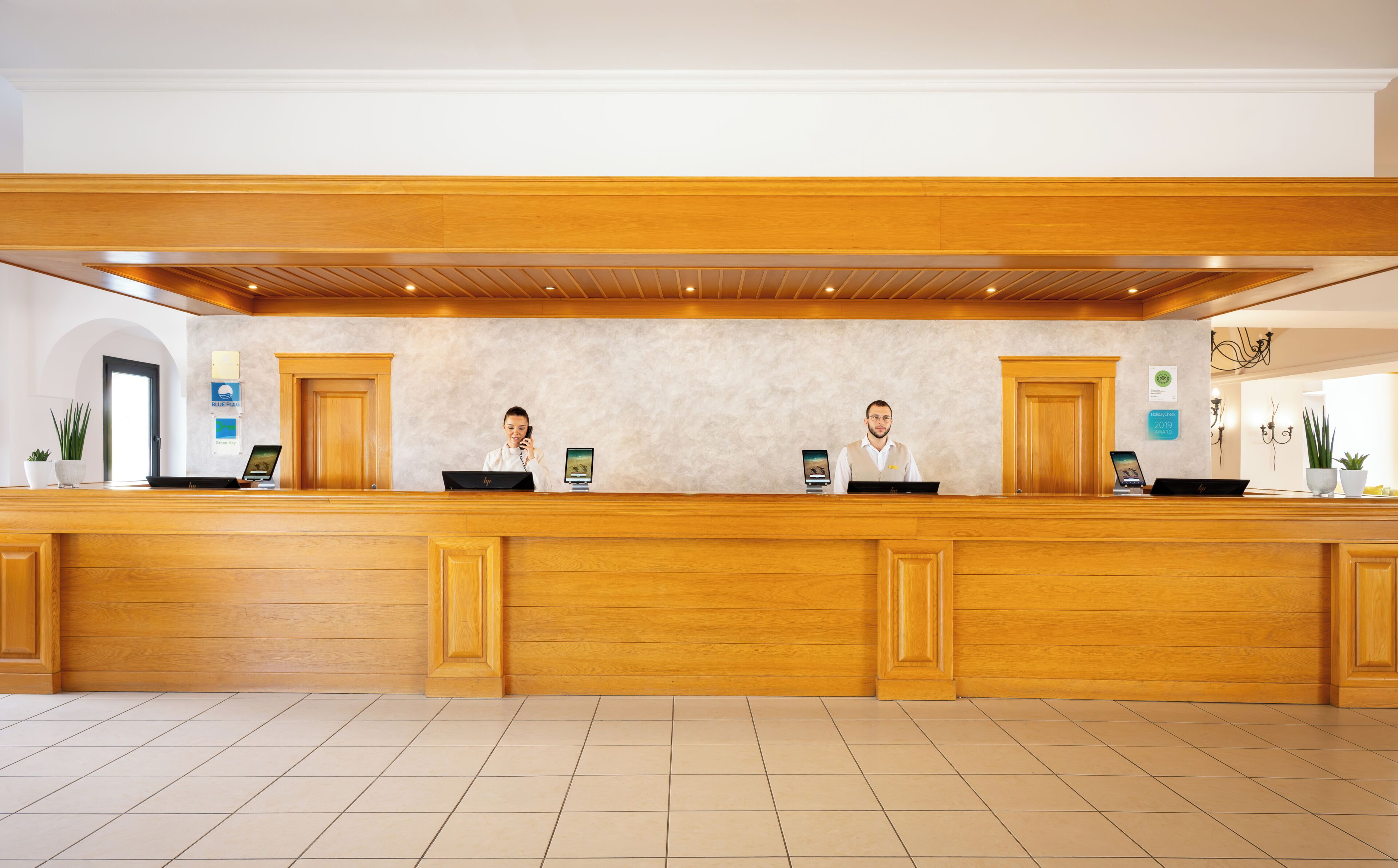 A reception desk with two employees, a wooden counter, and a tiled floor.