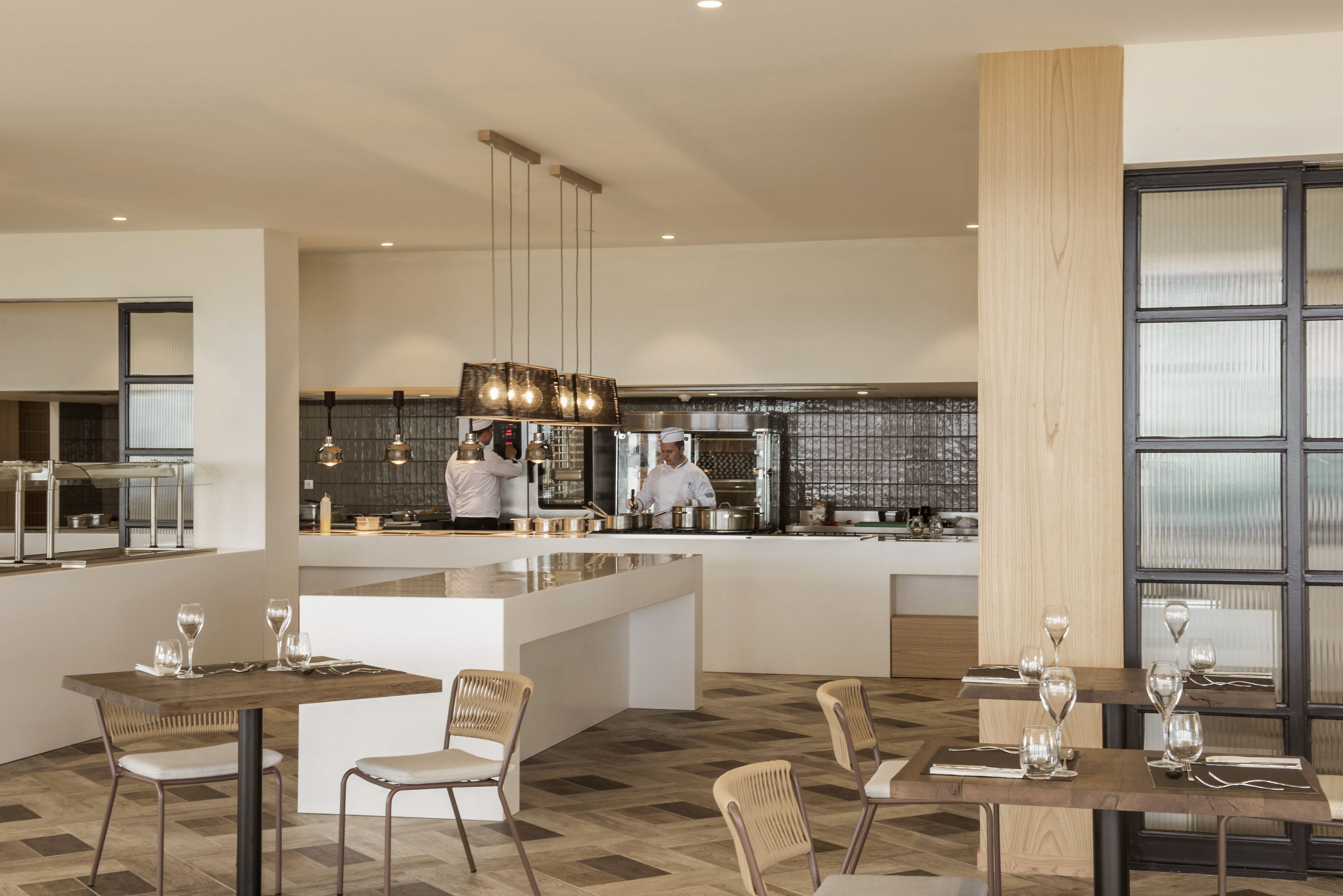 A modern restaurant with a central island, multiple dining tables, and a kitchen area in the background.