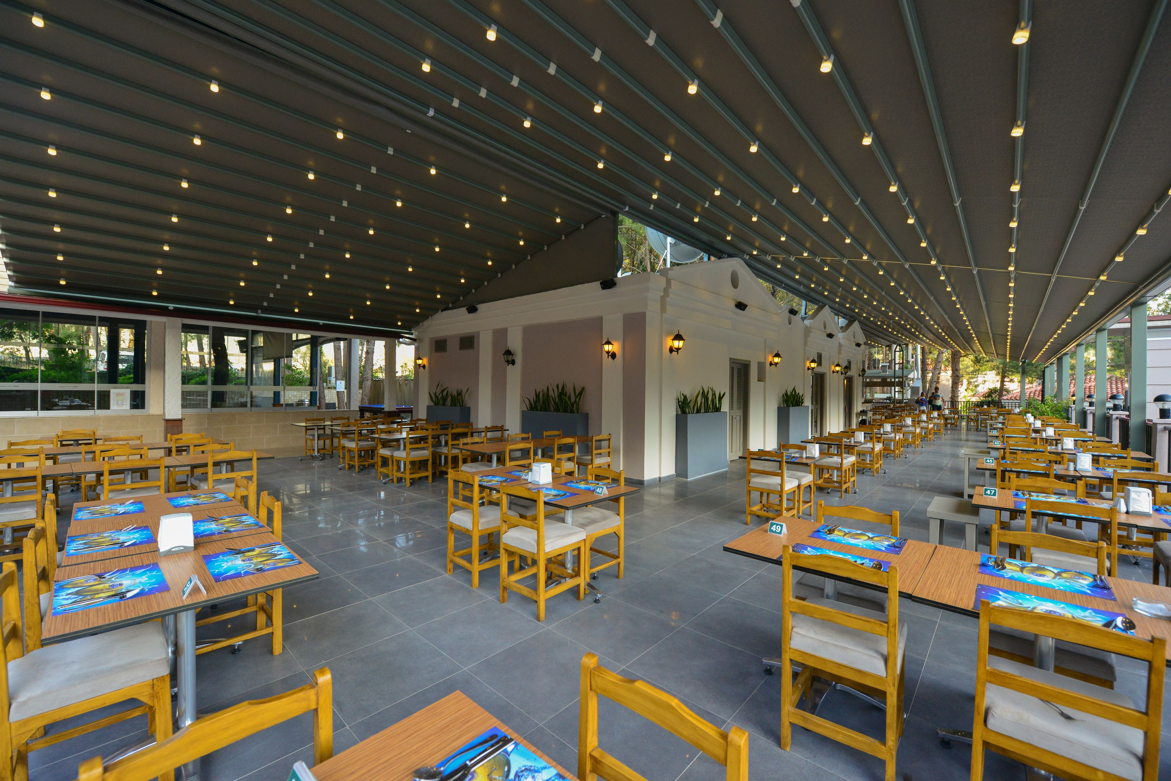 A spacious outdoor dining area with multiple tables and chairs, covered by a roof with evenly spaced lights.