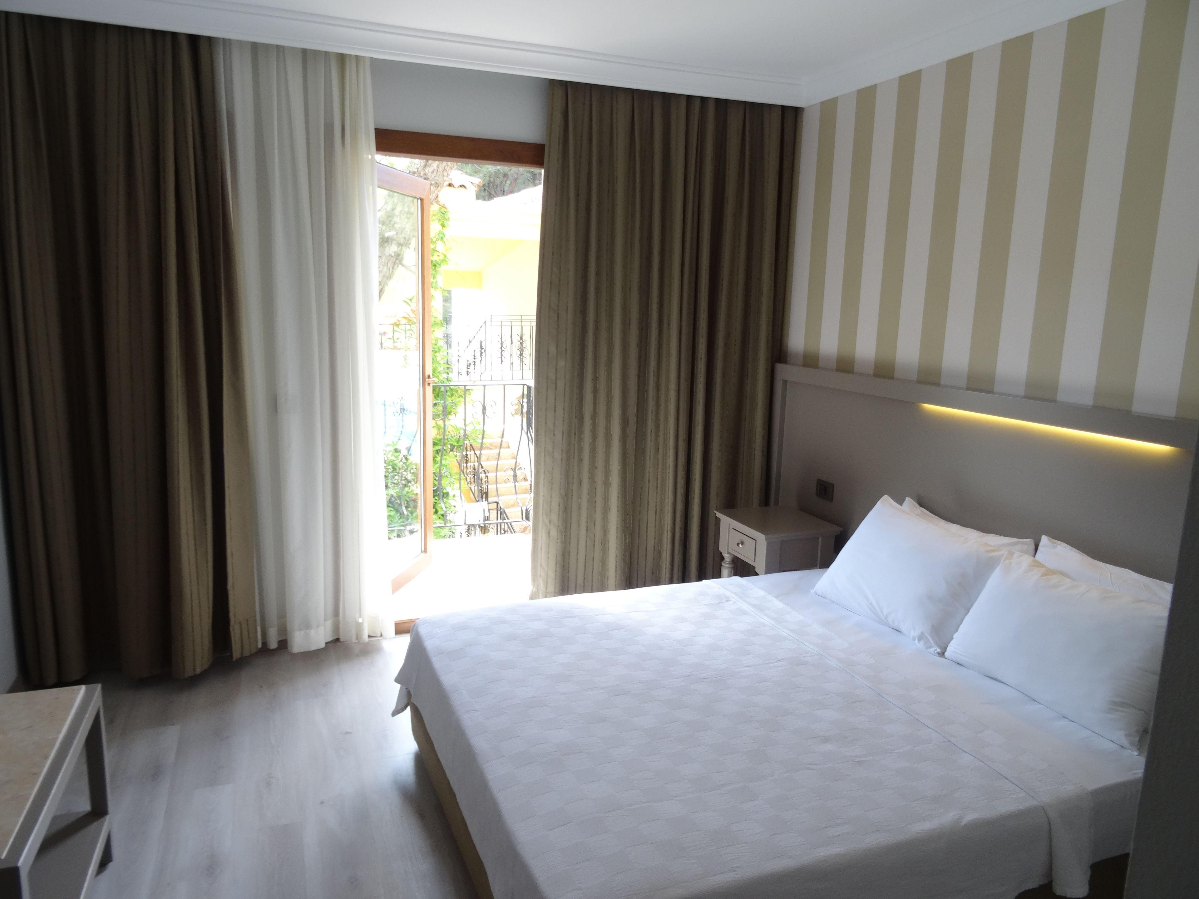 A hotel room with a bed, a nightstand, and a view of the outdoors.