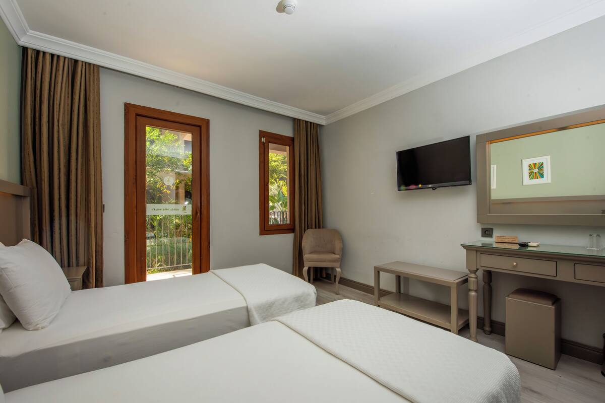 Habitación de hotel con dos camas individuales, un escritorio, una silla, un televisor y una ventana con cortinas.