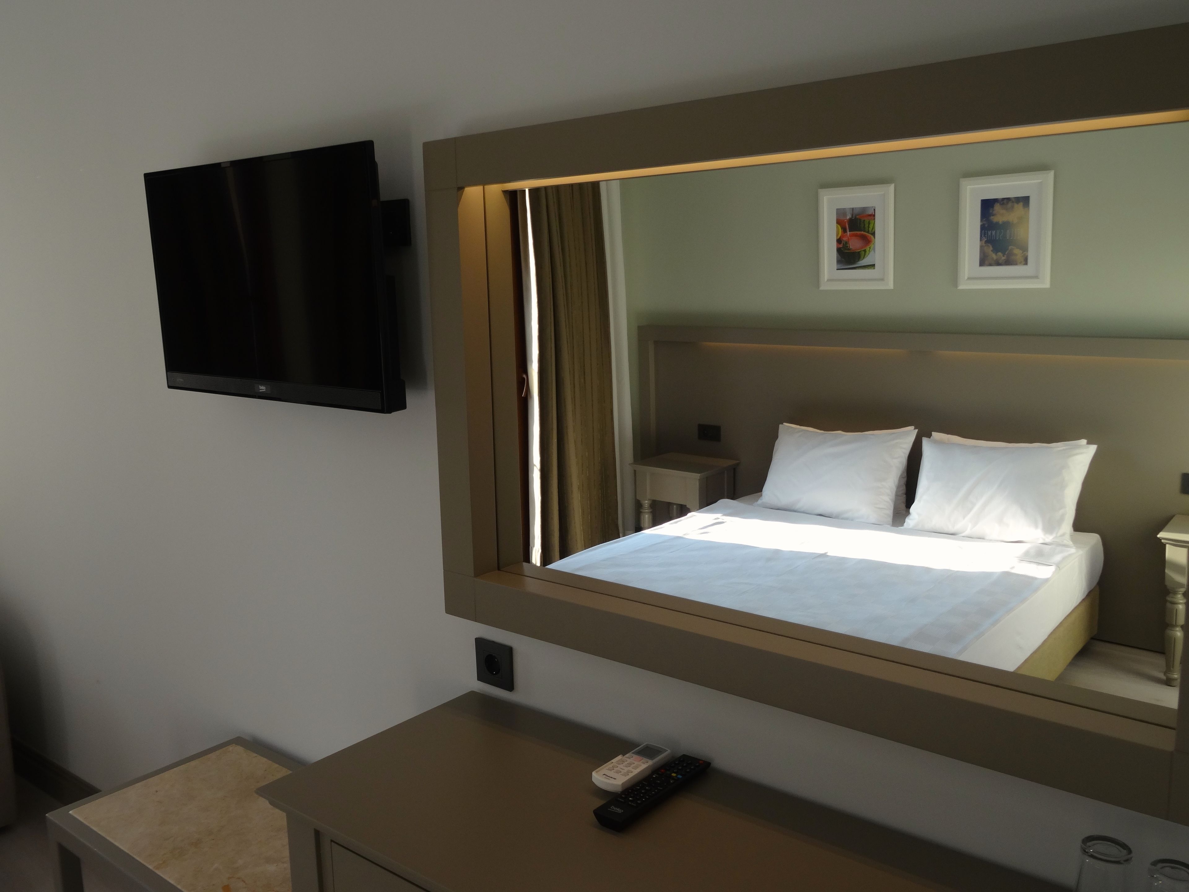 A hotel room with a large bed, a bedside table with a remote control, a flat-screen TV mounted on the wall, and a small nightstand with a lamp.