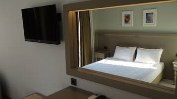 A hotel room with a large bed, a bedside table with a remote control, a flat-screen TV mounted on the wall, and a small nightstand with a lamp.