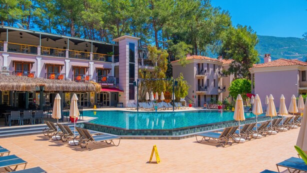 A pool area with lounge chairs and umbrellas, surrounded by buildings with balconies and a mountain in the background. - Hotel Pine Valley (Fethiye)