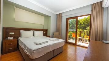 A hotel room with a large bed, a wooden nightstand, a chair, and a balcony with a view of greenery.